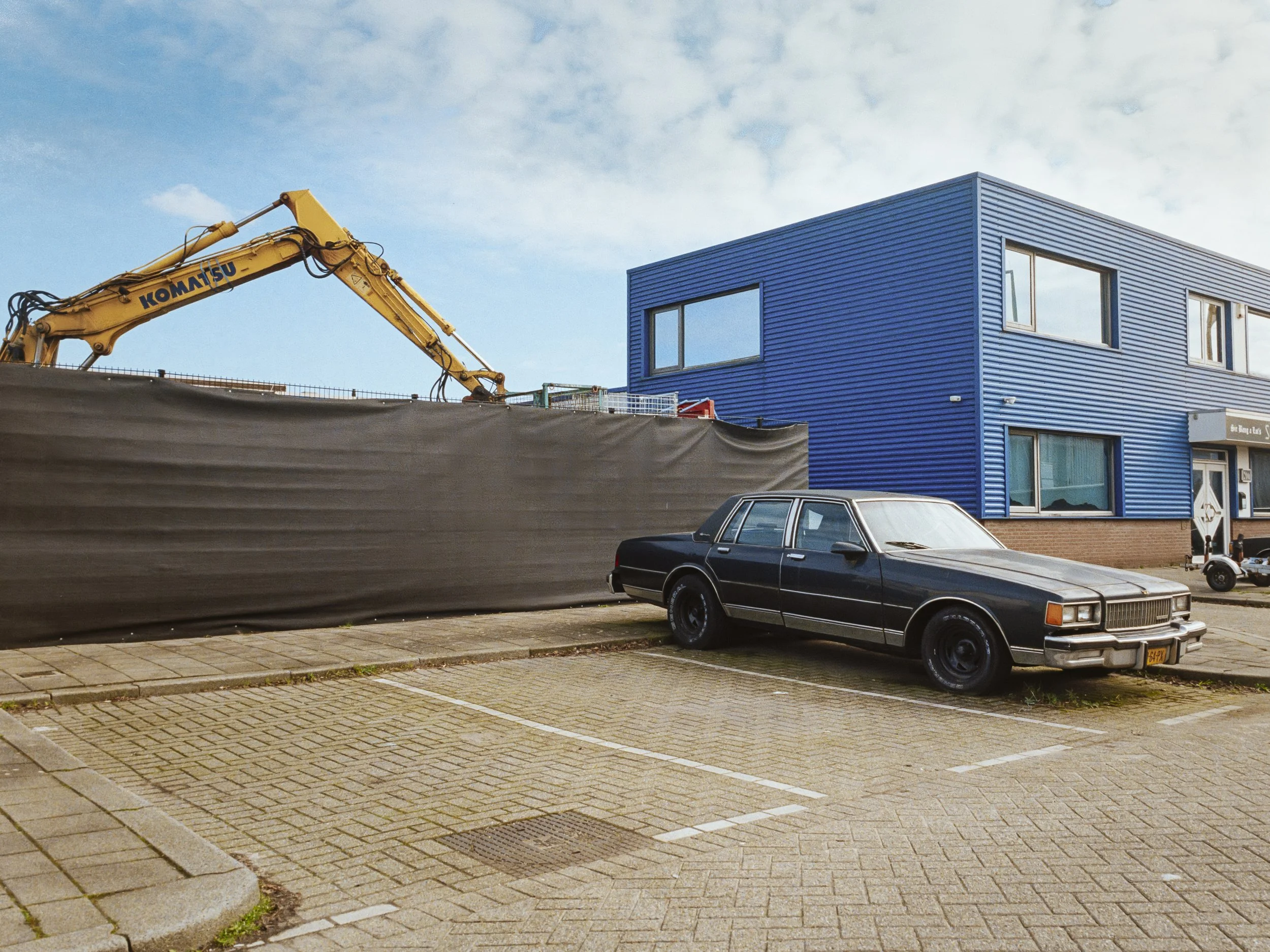 A parking lot with designated spaces, a black vintage car, a construction excavator with a yellow arm, a modern blue building with large windows, a street lamp, and a clear sky with some clouds.