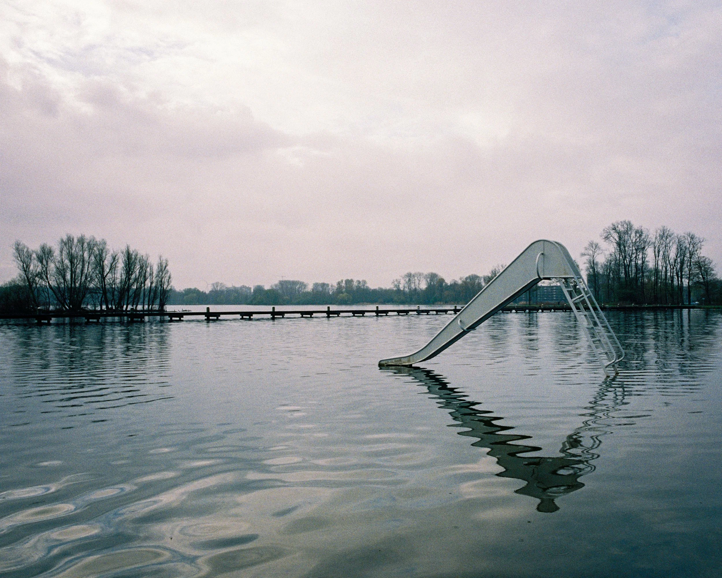 A lakeside view with a dock and a metal diving board, leafless trees along the shoreline, and overcast sky.