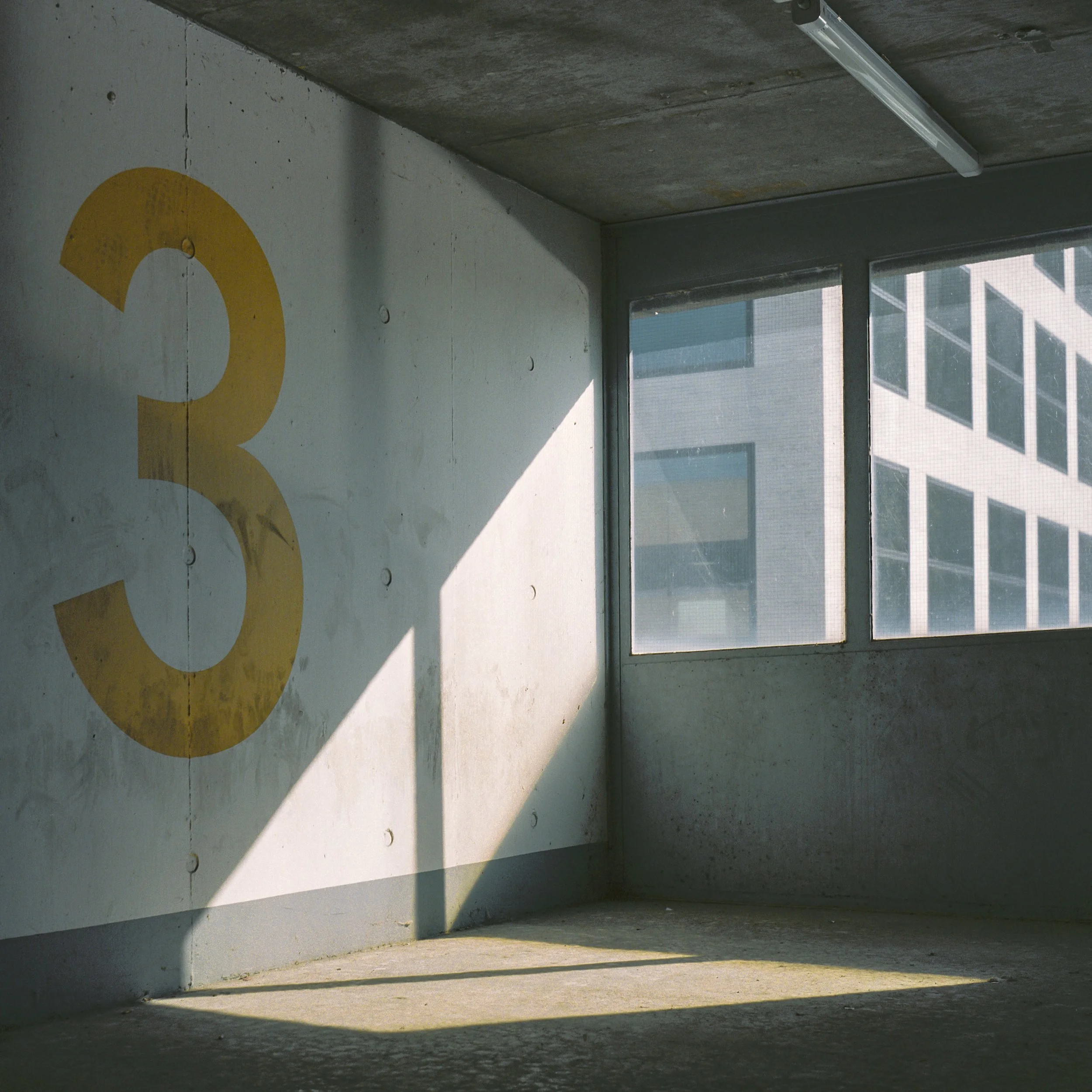 Concrete interior space with a window showing a building outside, a fluorescent light fixture on the ceiling, and a wall with partially visible yellow painted letters.