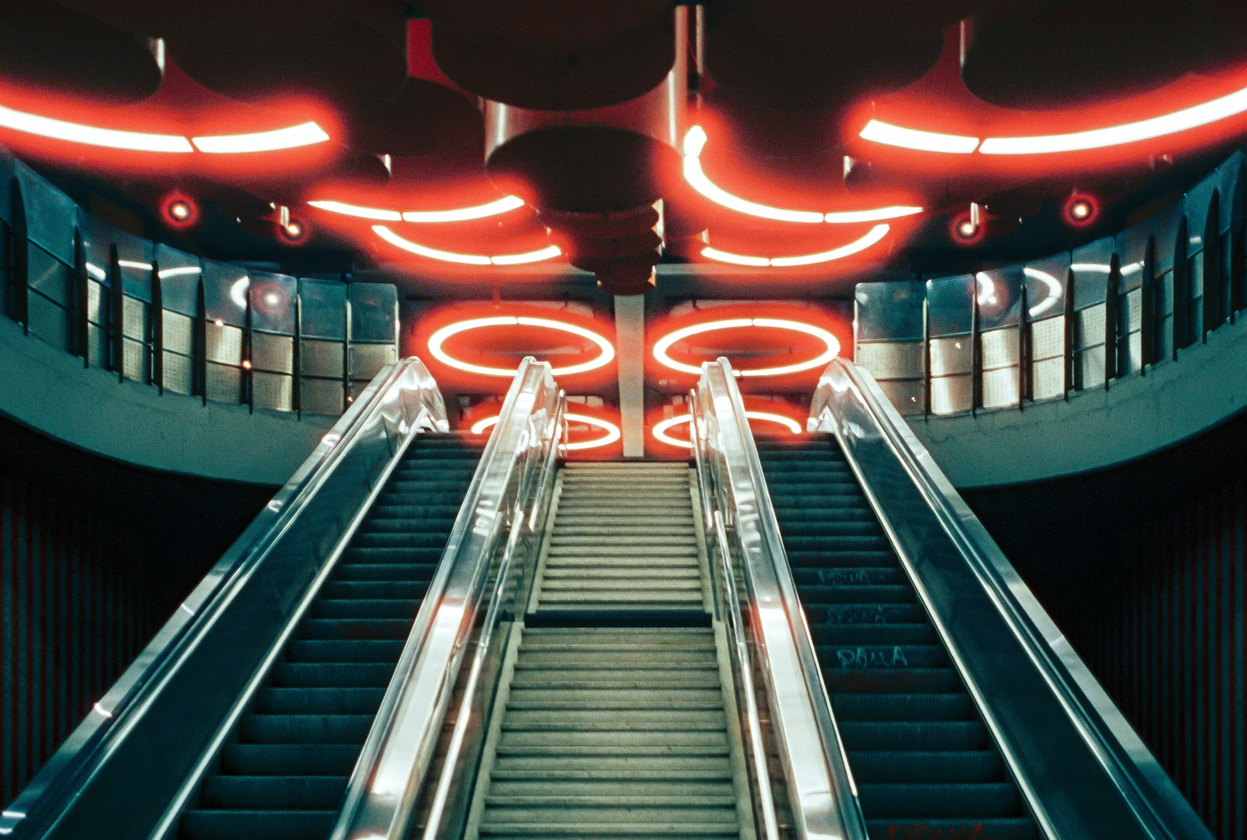 Escalator leading to a circular, neon-lit ceiling with red and white lighting accents in a modern building.