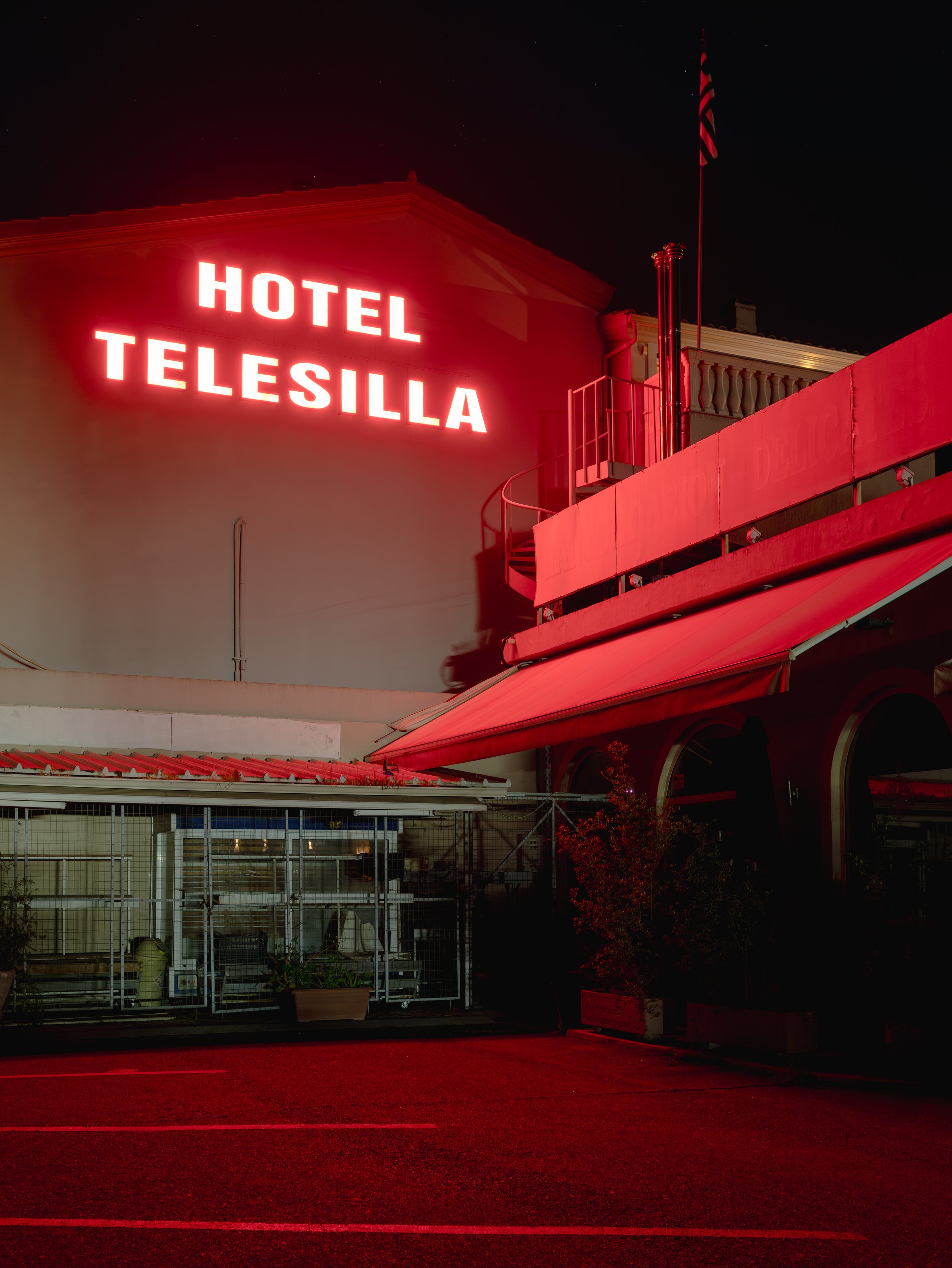 Night view of a building with a bright red neon sign reading 'HOTEL TELESILLA', a flagpole with an American flag, and a dark parking lot in the foreground.