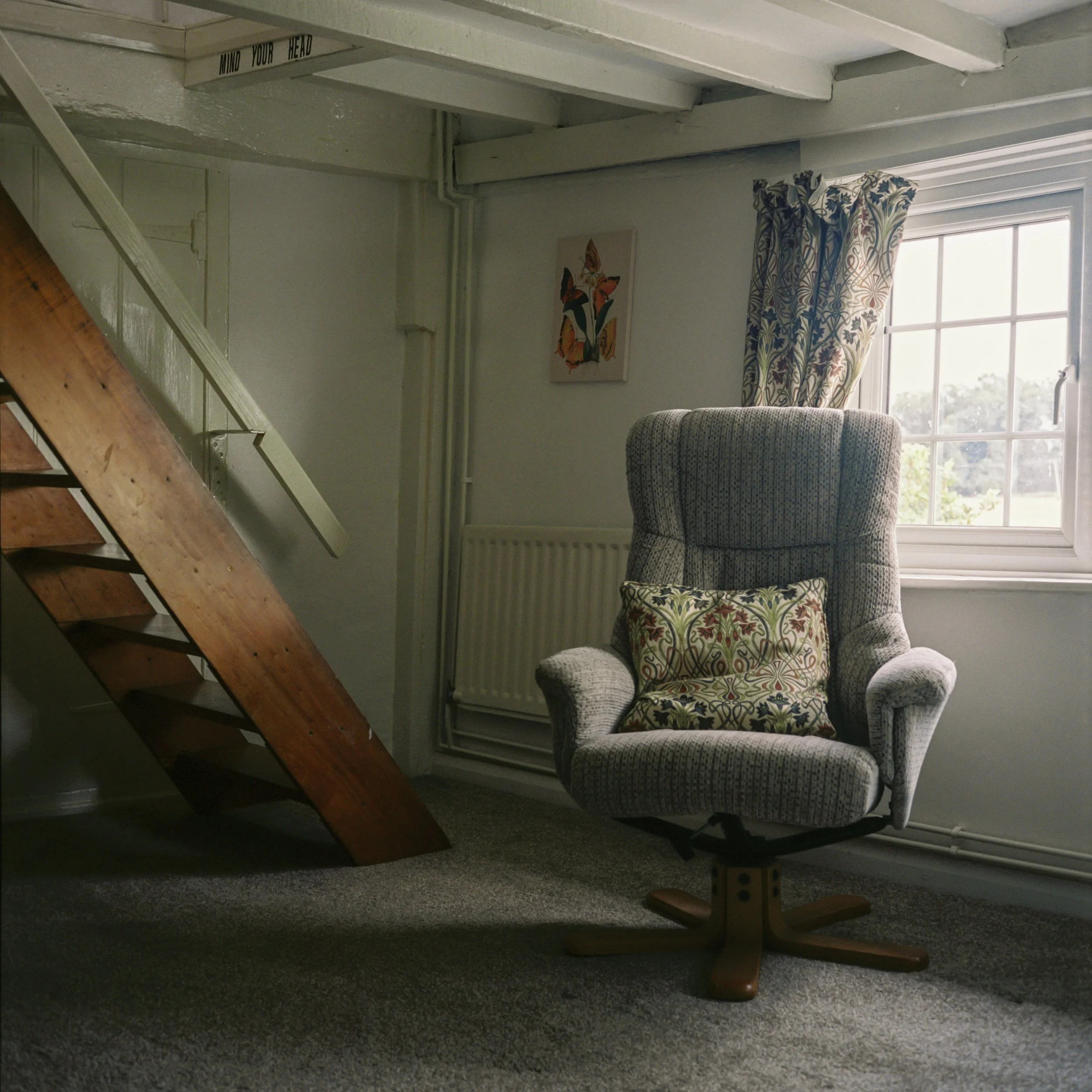 View of a cozy living room corner with a patterned armchair, a window with floral curtains, a radiator, and a framed butterfly art print on the wall.