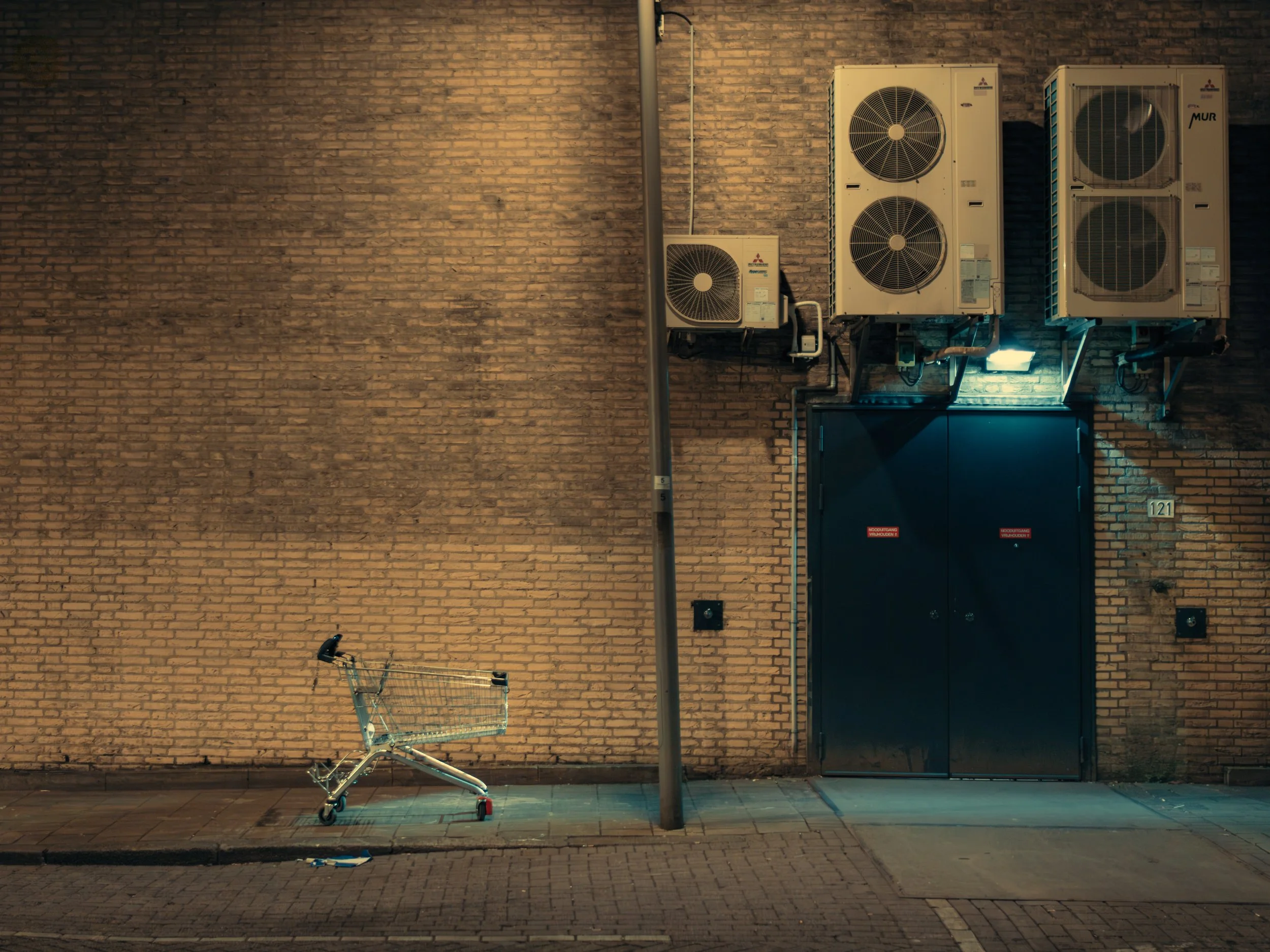 An empty shopping cart tipped over on a sidewalk in front of a brick wall. A street lamp and air conditioning units are mounted on the wall. A small sign with number 121 is visible.