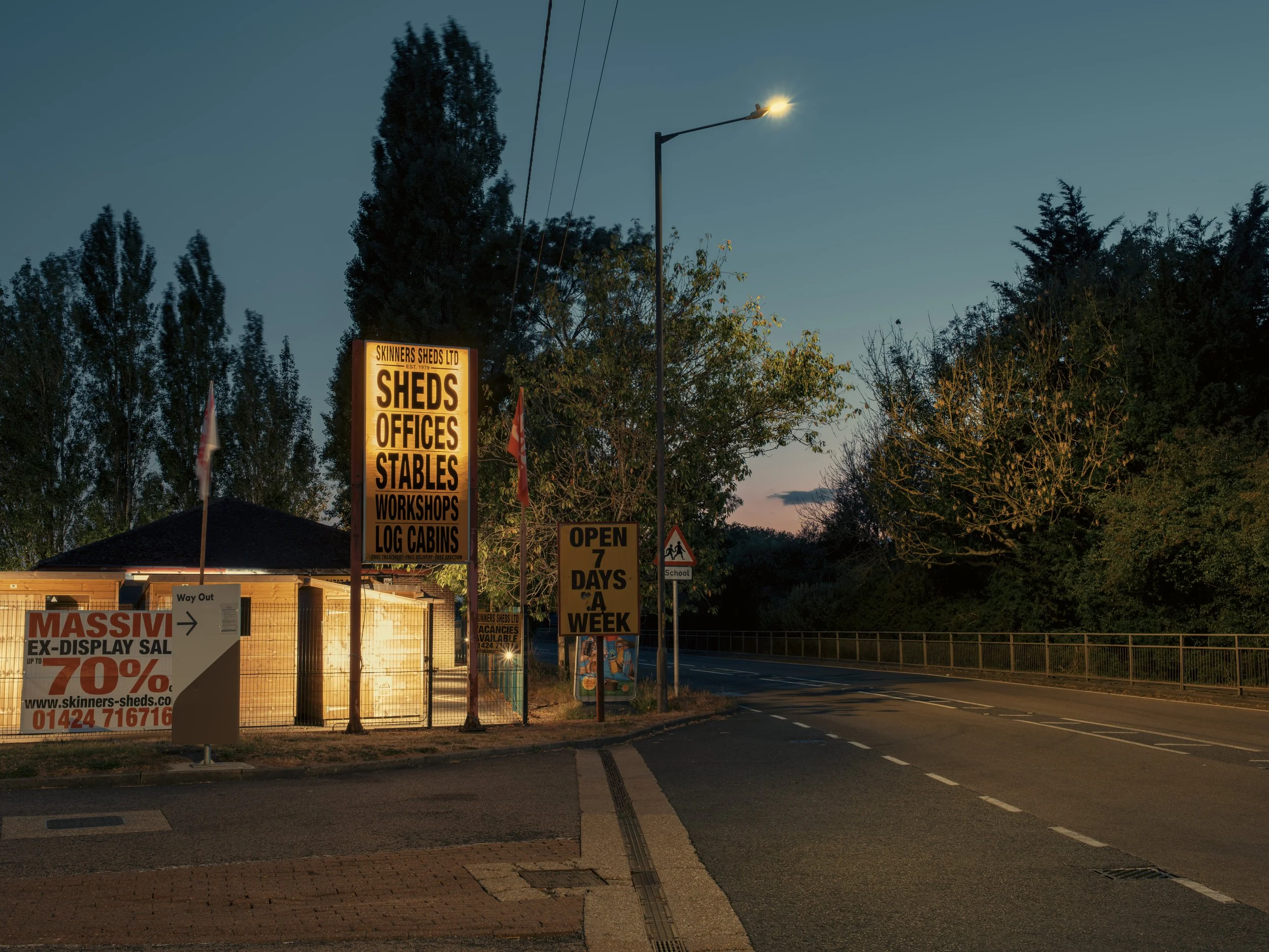 A roadside area with several signs advertising sheds, offices, stables, workshops, and log cabins for sale. A streetlight is turned on, illuminating the signs and trees as dusk approaches.