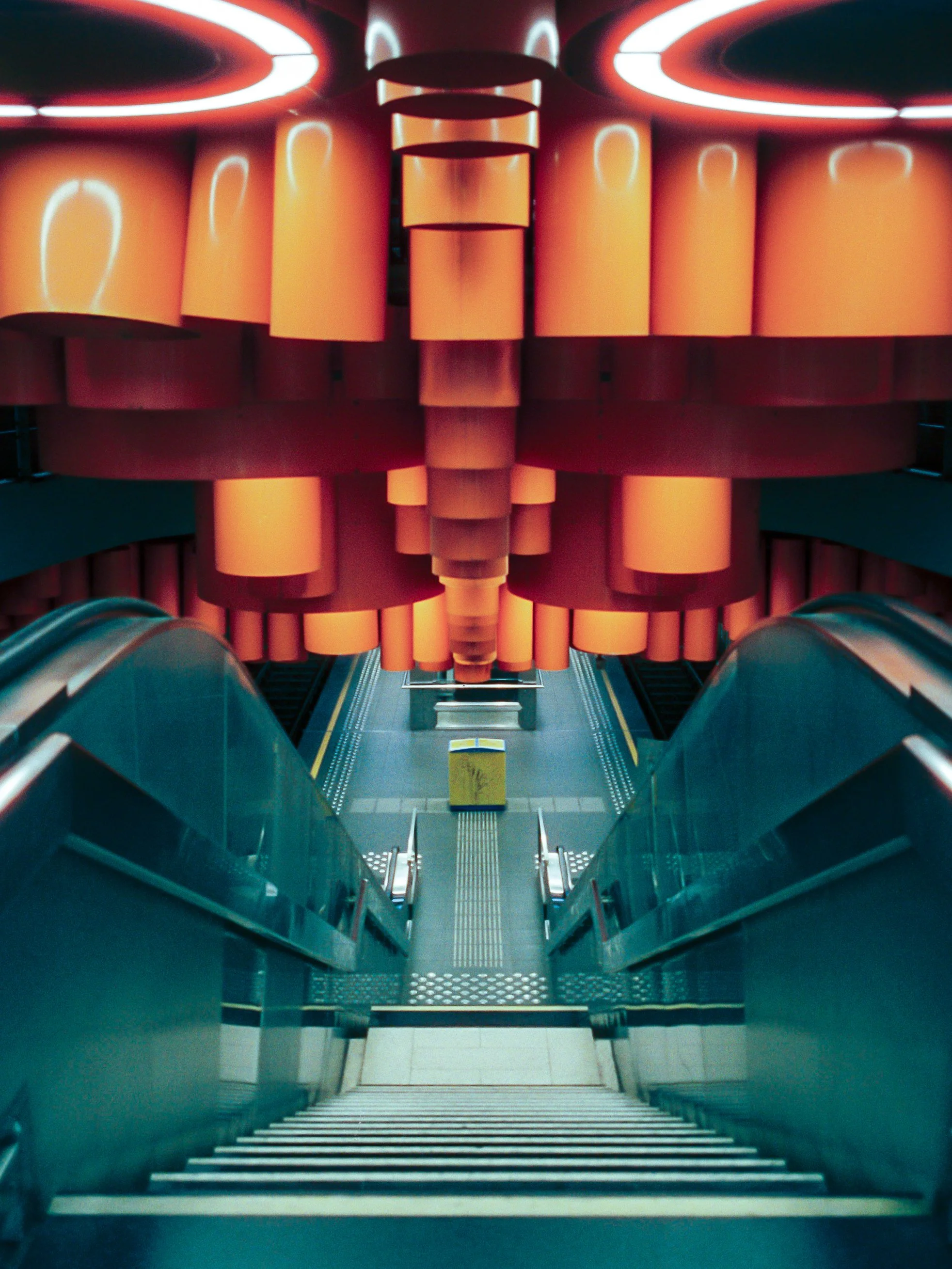 Inside view of an escalator leading down, with bright orange cylindrical structures and a metal grate area in the center, illuminated by circular lights on the ceiling.