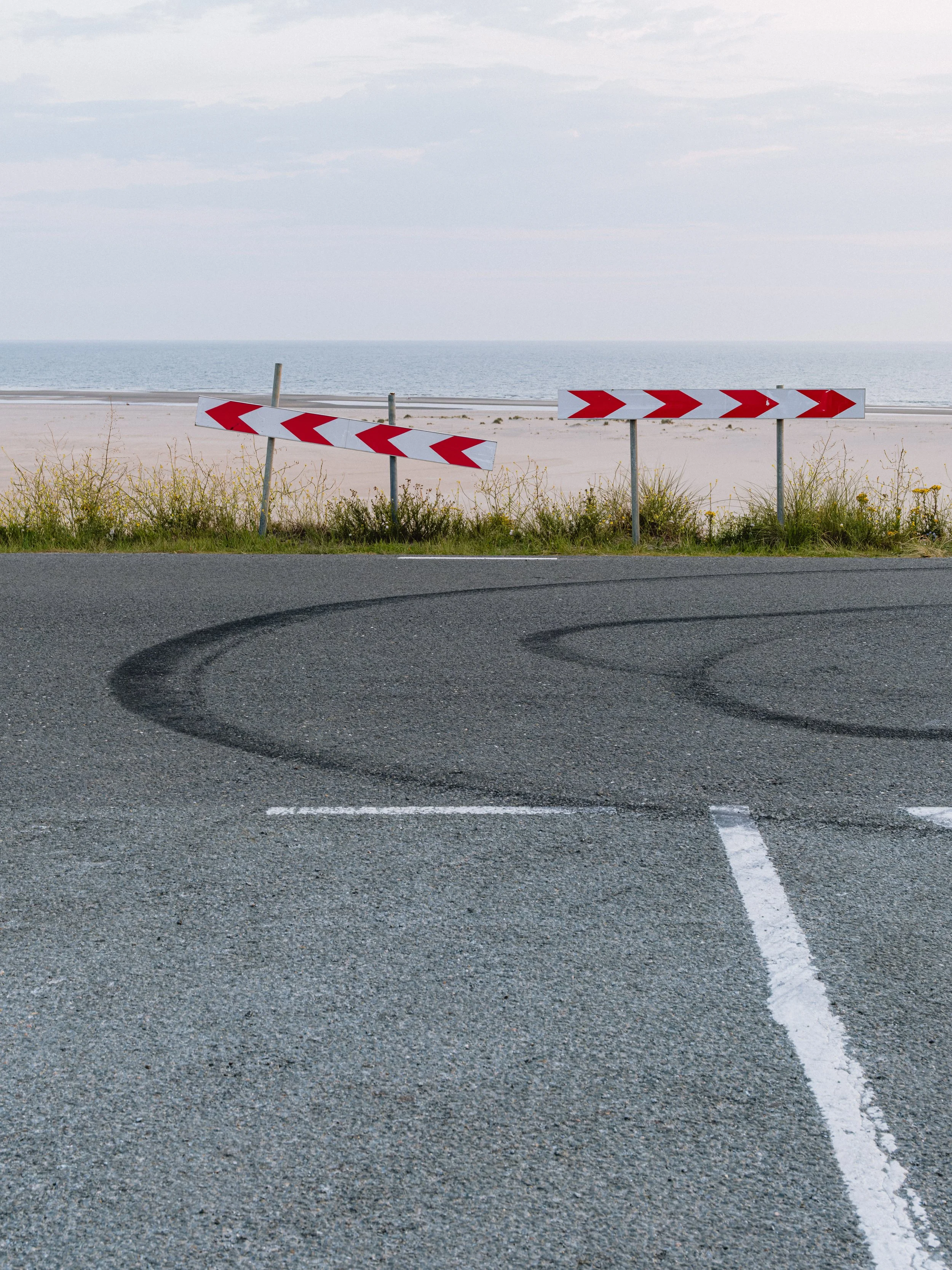 A road with a tire skid mark, a white dashed line, and red and white chevron road signs near a grassy area by a beach with sand and water in the background.