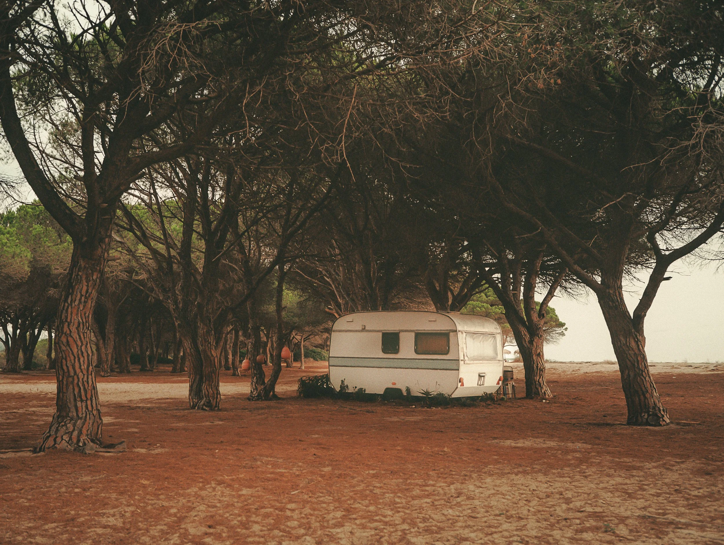 A white camper trailer parked on a patch of dirt under trees with leafless branches, next to a chair, near a beach or lake.