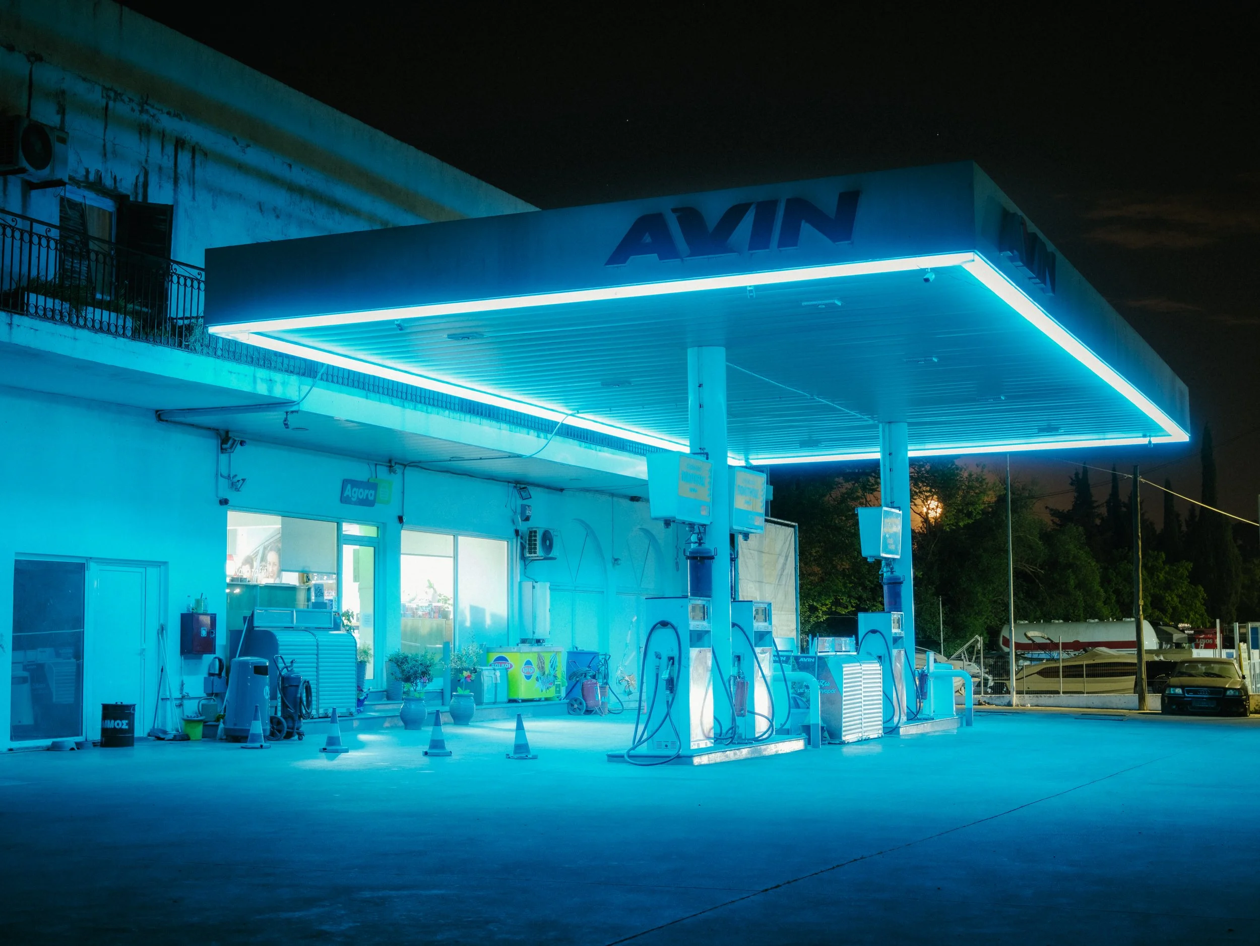 Nighttime of a gasoline station with blue lighting, multiple fuel pumps, and an AYM sign on the canopy. The station appears to be closed or not in use, with no cars or people present.