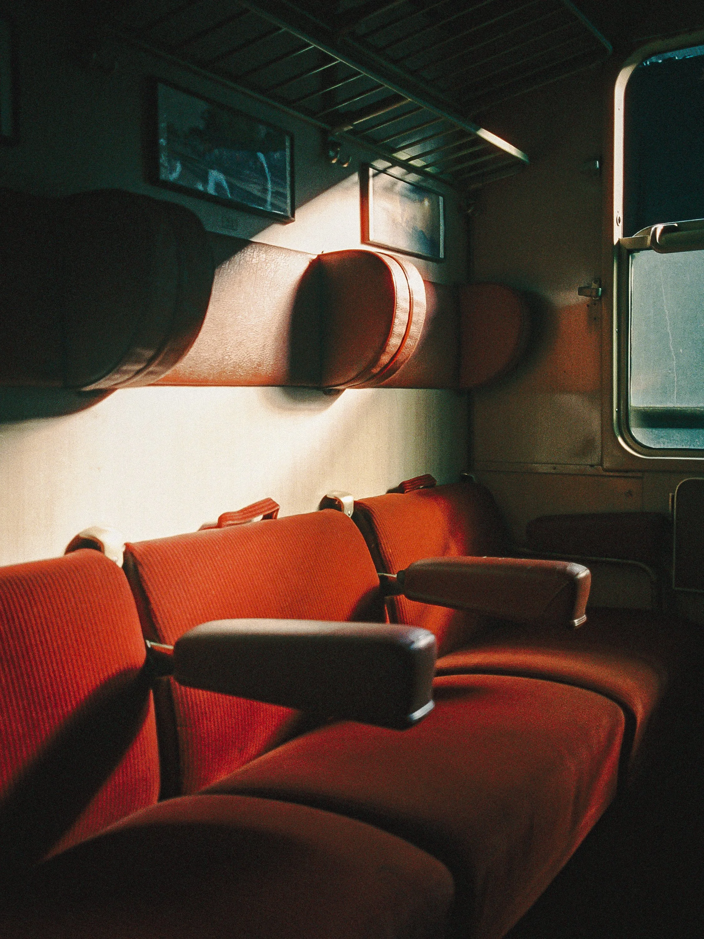 Interior of a bus with red cushioned seats, some with black armrests, and two television screens on the wall at the front. Sunlight is casting shadows on the seats and walls.