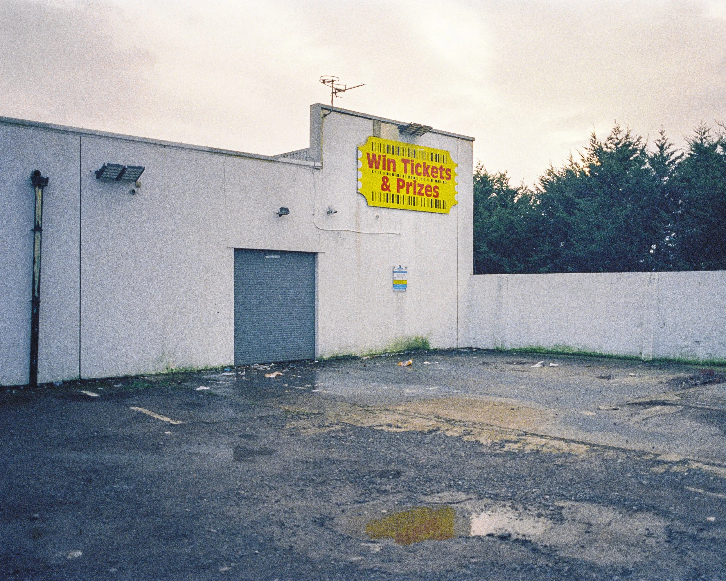 An empty parking lot with a white building featuring a large yellow sign reading 'Win Tickets & Prizes' and a gray roller door. There are some small debris on the dark pavement, and a row of trees is visible in the background under a cloudy sky.