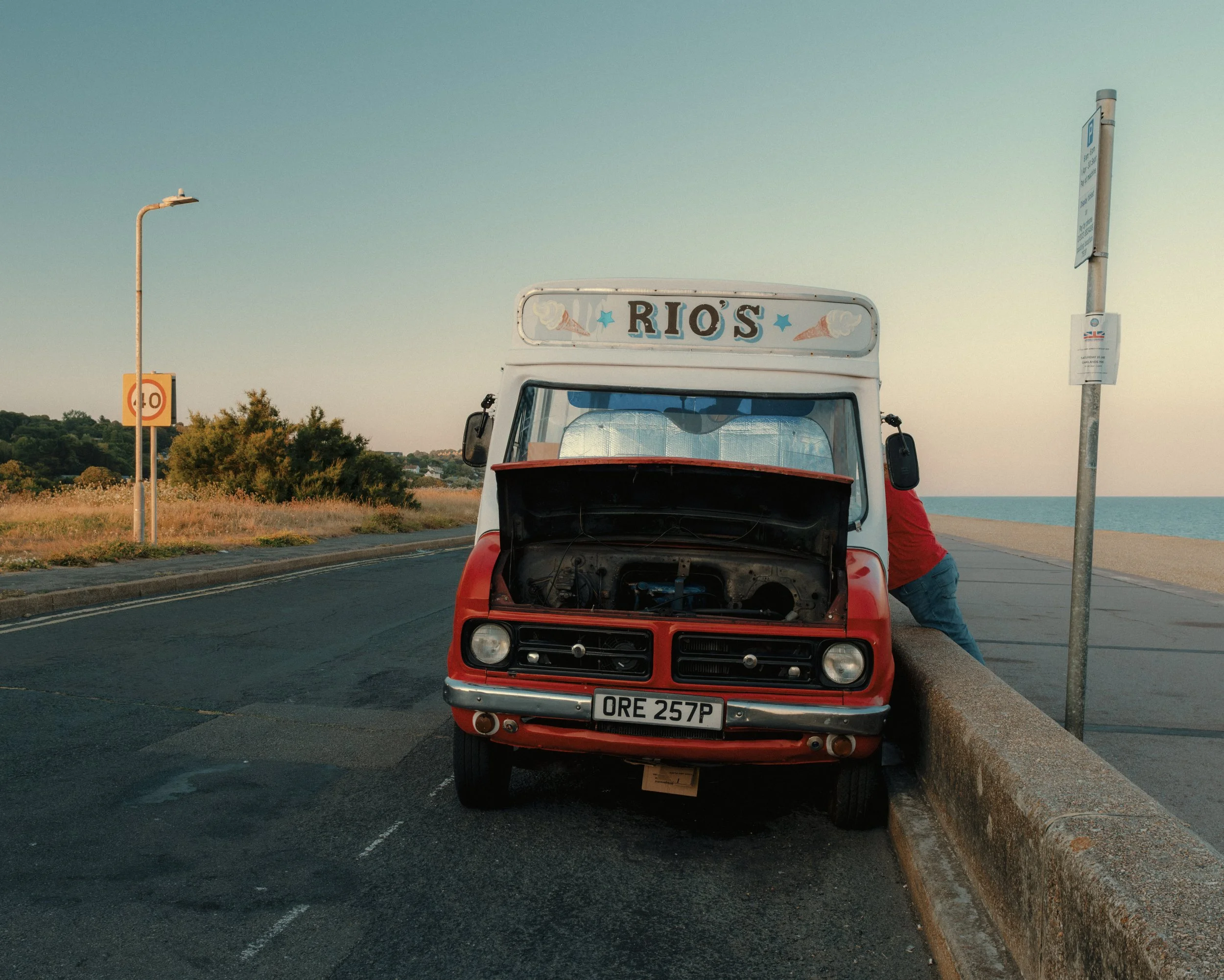 Old red car with its hood open parked along a roadside, with a person working on it. There are two streetlights and a bus stop sign nearby, and a landscape with trees and water in the background during sunset or sunrise.