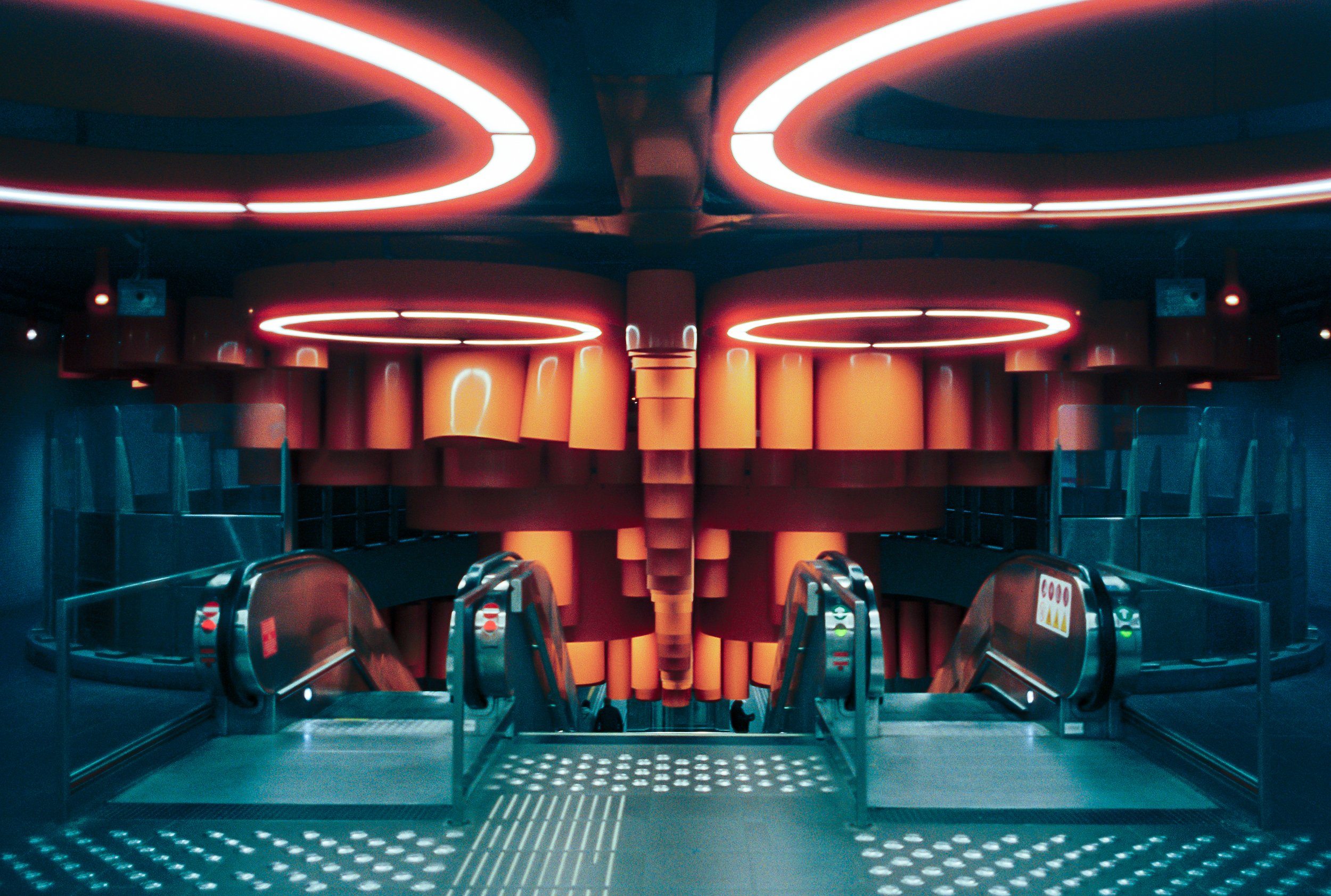 Inside an airport, view of an escalator with neon lighting overhead, leading up to a walkway with orange pillars and a curved railing.