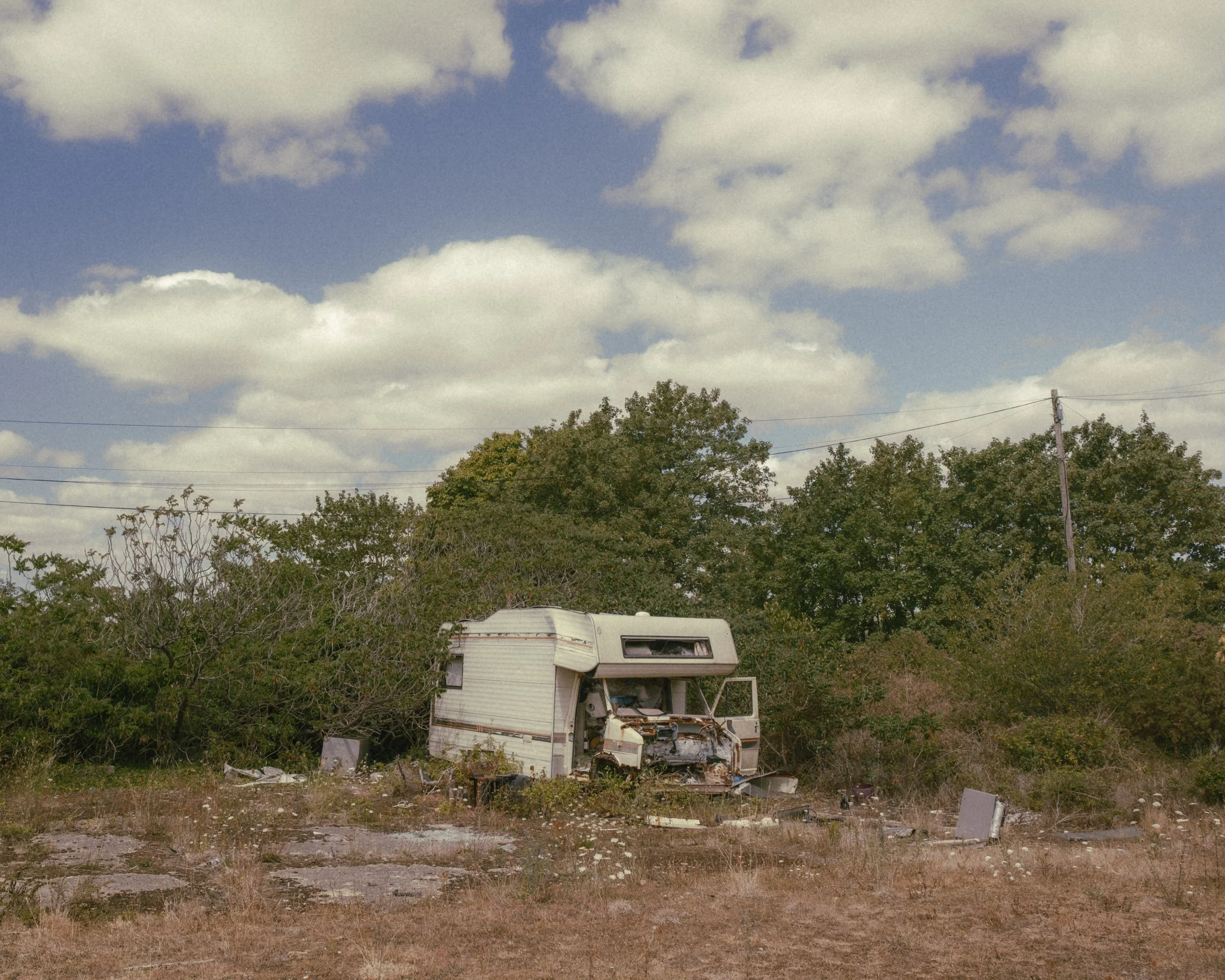 A damaged camper trailer with missing parts in a grassy area, with trees and power lines in the background under a partly cloudy sky.
