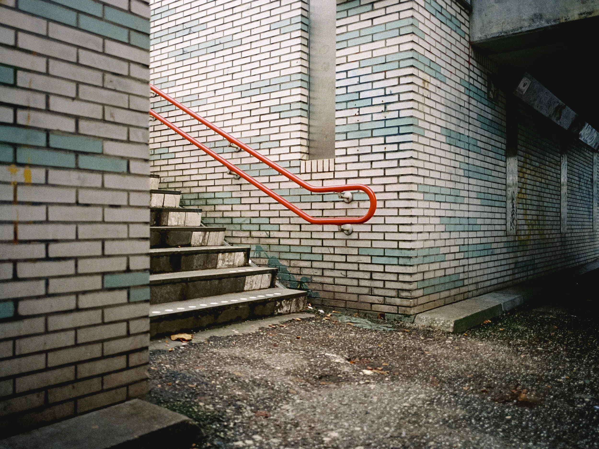 A brick exterior with a staircase and a red handrail leading upward. There is a small gap between bricks and some leaves and debris on the ground.