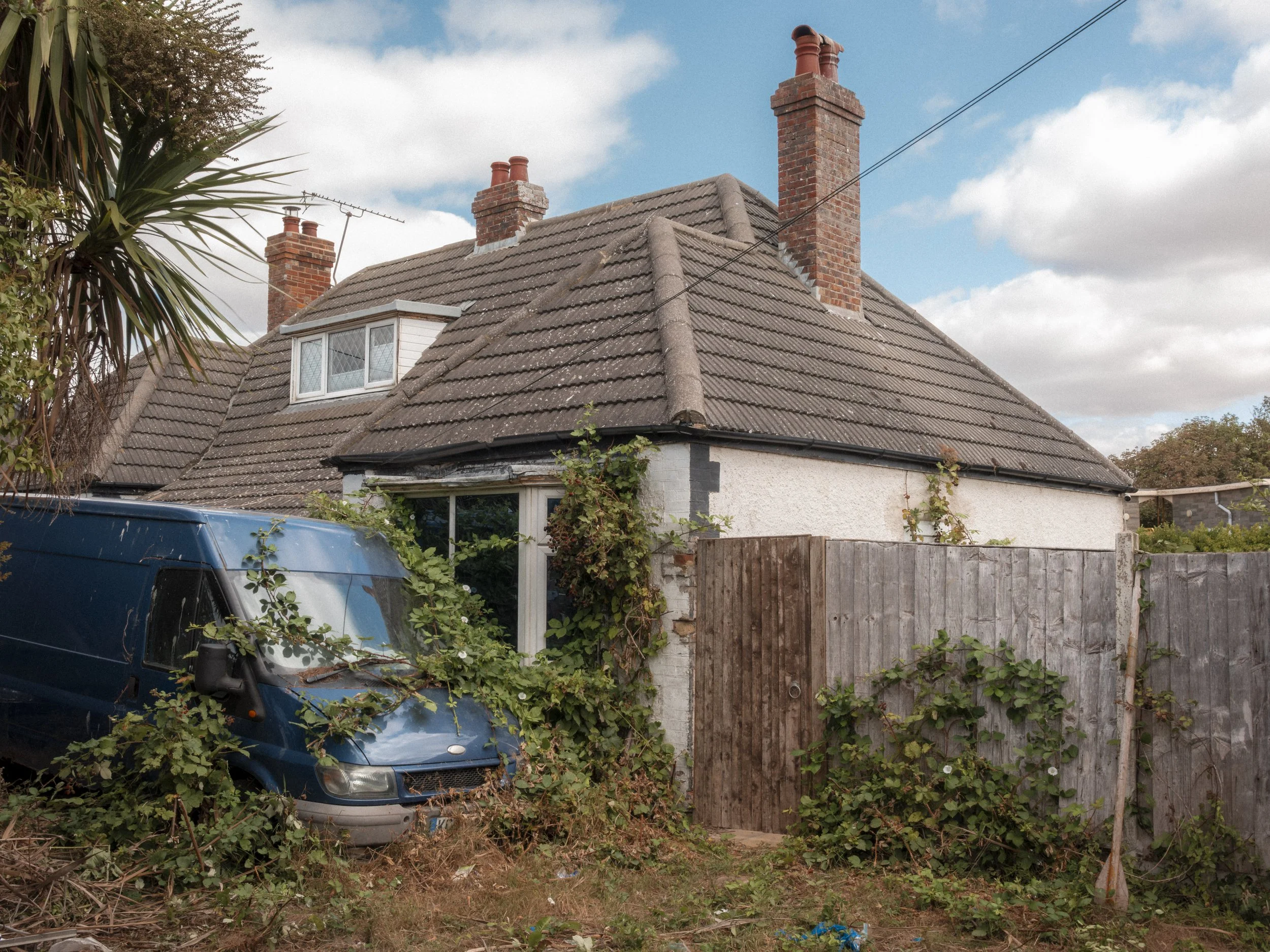 A house with a sloped tiled roof, three red brick chimneys, and a small attic window. There are trees and bushes surrounding the house, and an overgrown yard with a blue vehicle partially visible in front.