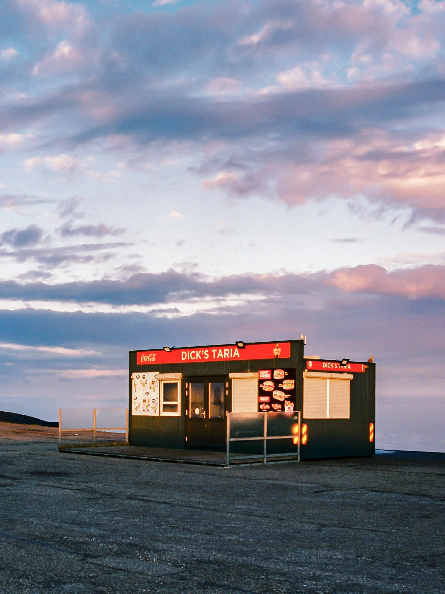 A fast-food kiosk in an empty parking lot under a cloudy sky at sunset, with a sign reading 'Dick's Taria' and menu images displayed on the side.