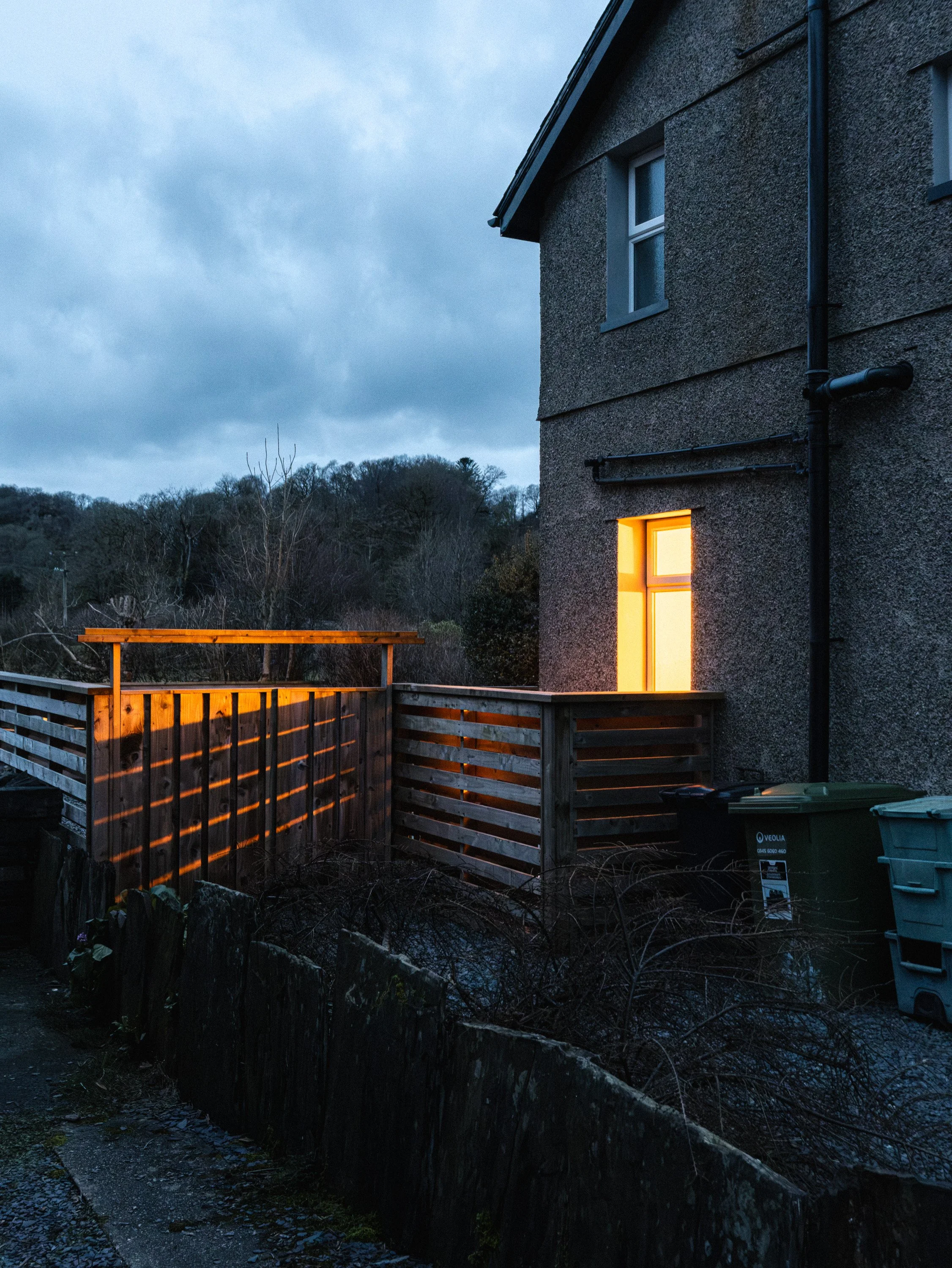 A house with a lit window at dusk, surrounded by barren trees and a wooden fence, under a cloudy sky.