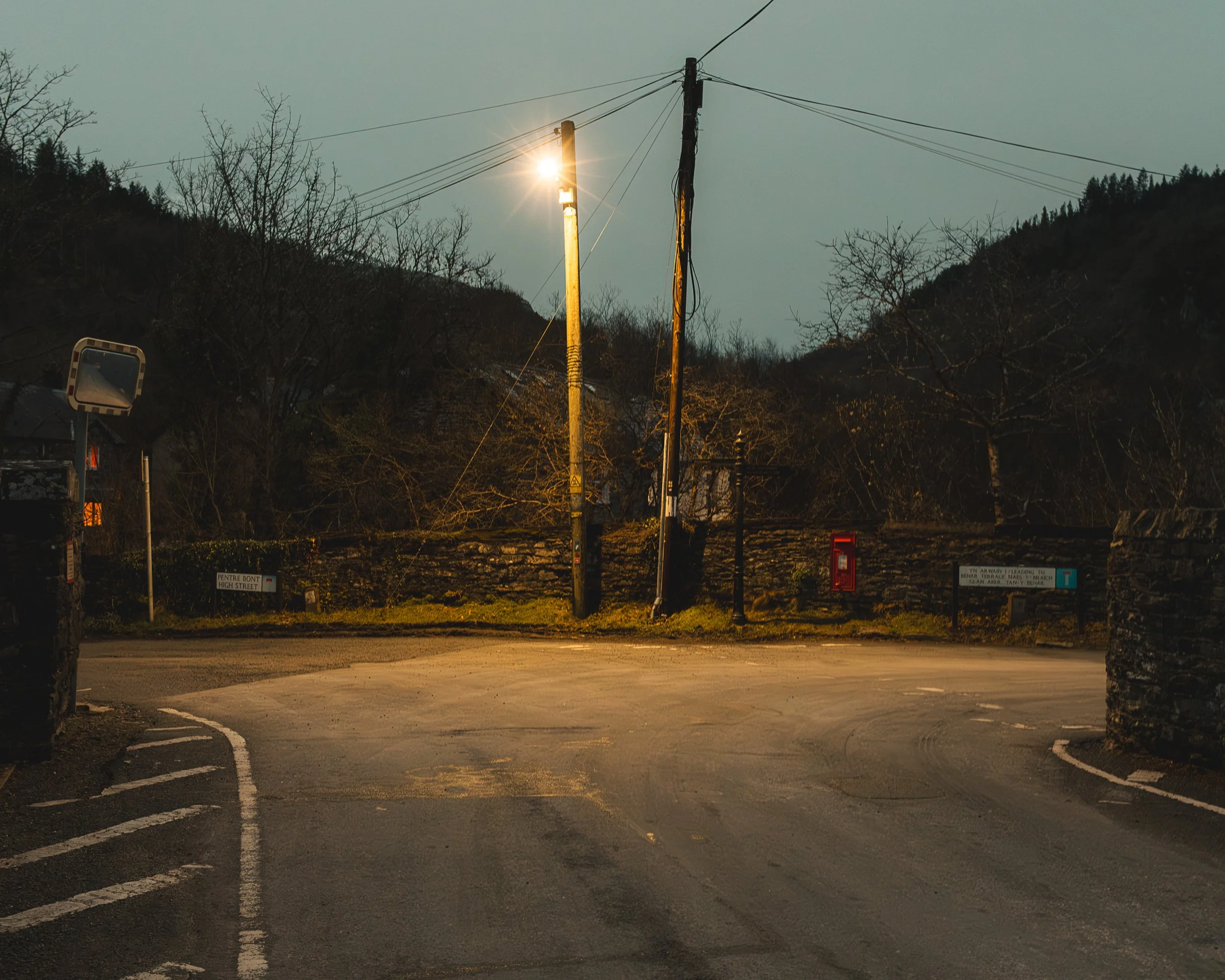 Nighttime view of a street with a utility pole and wires illuminated by a streetlight, trees and mountains in the background, and a mirror reflecting the sky.