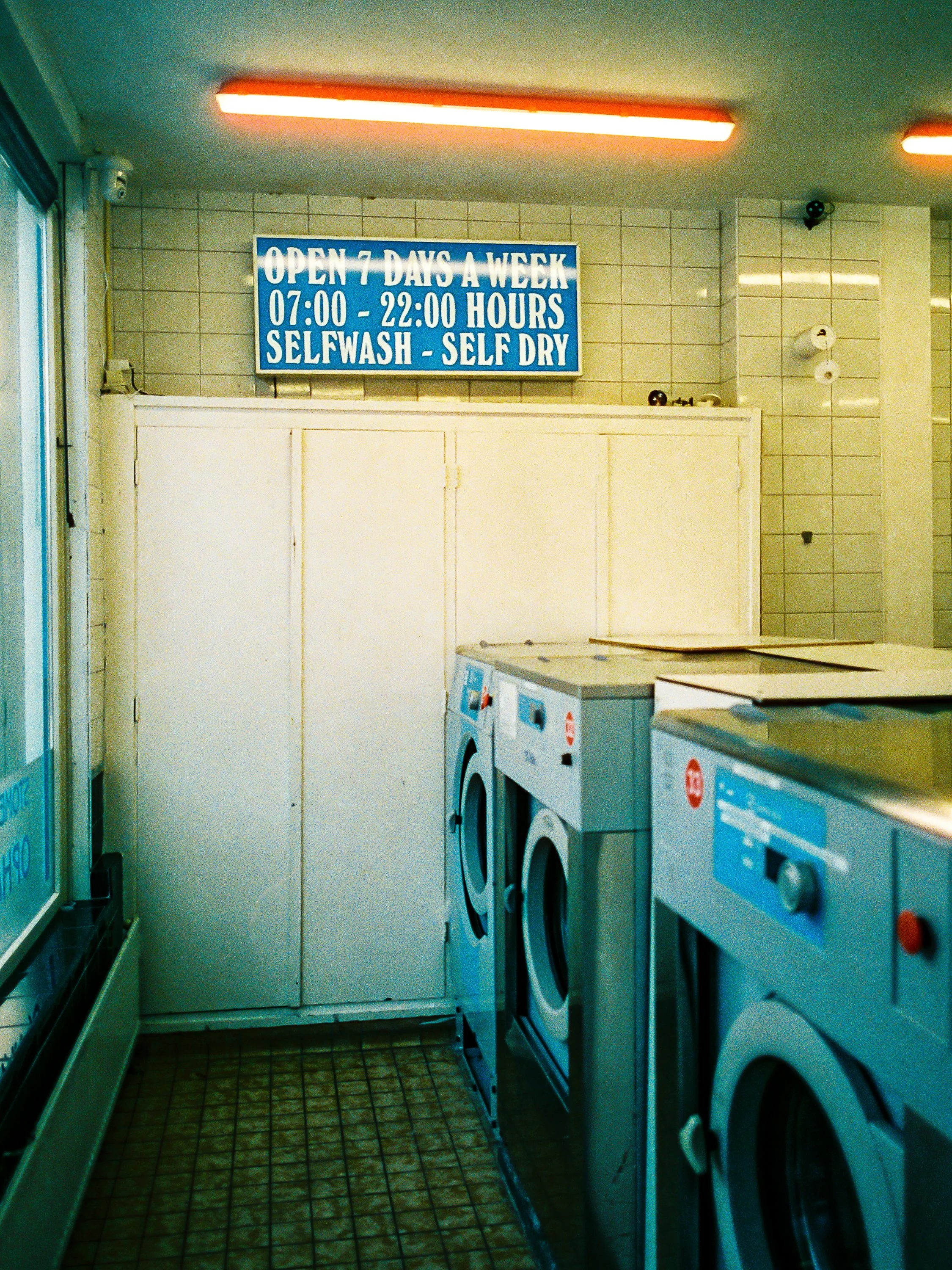 Inside a laundromat with washing machines along the wall and a sign indicating it is open 7 days a week from 7:00 to 10:00 PM, featuring self-wash and self-dry services.