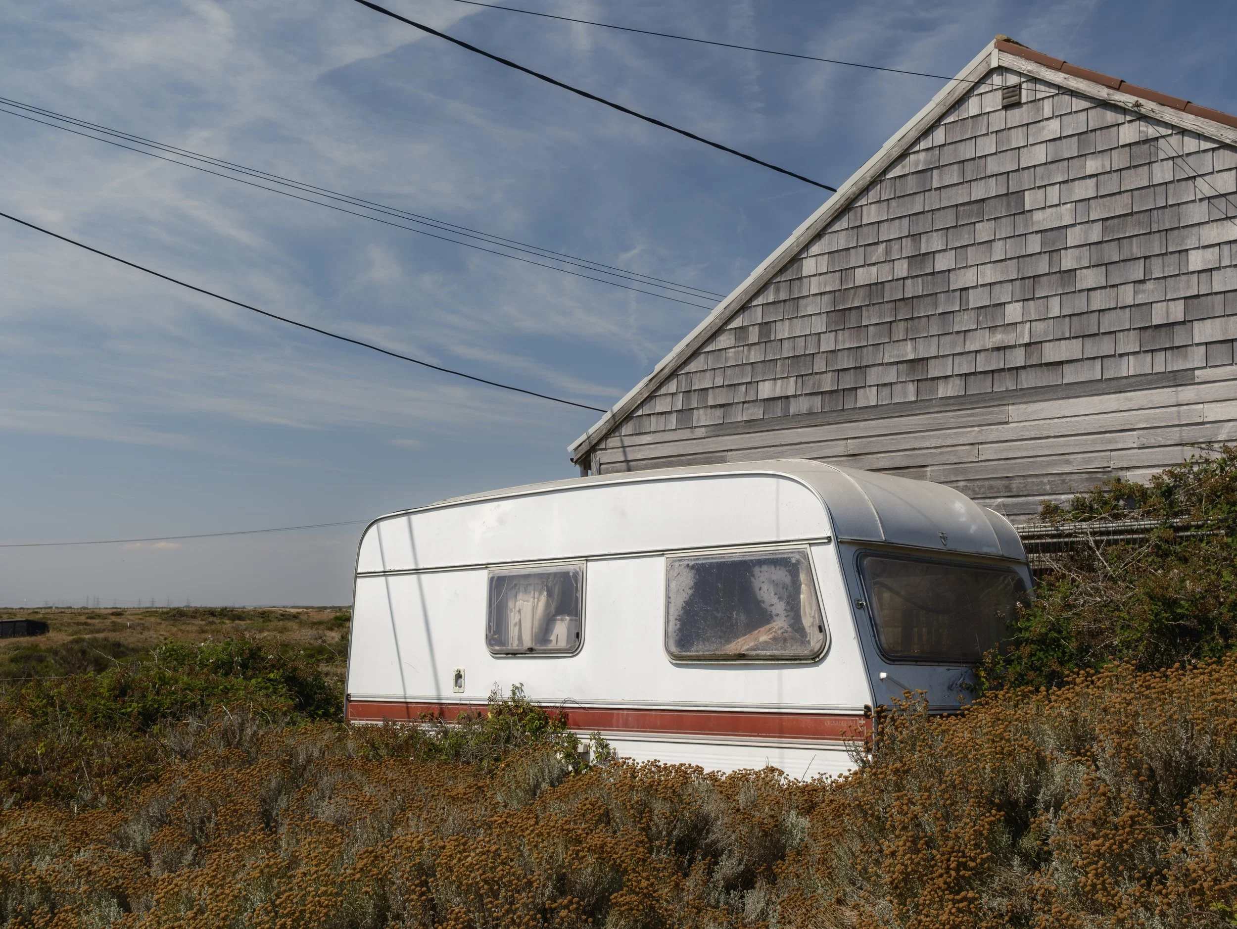 An old white camper trailer with red stripe parked in a grassy area with shrubs, next to a weathered wooden building with gray shingles, under a blue sky with wispy clouds.