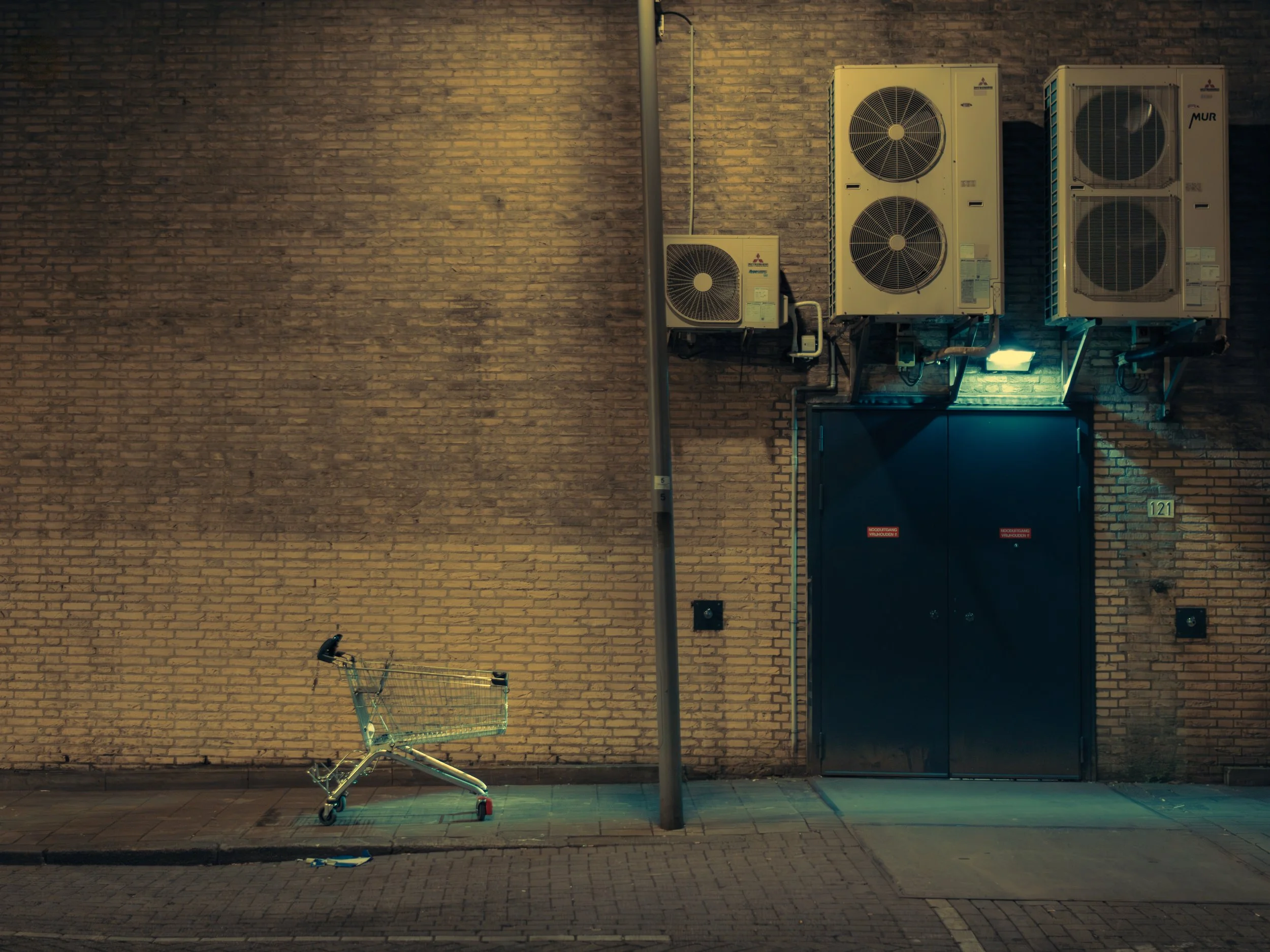 A shopping cart tipped over on the sidewalk in front of a brick wall, with a street lamp, air conditioning units, and a loading dock door nearby.
