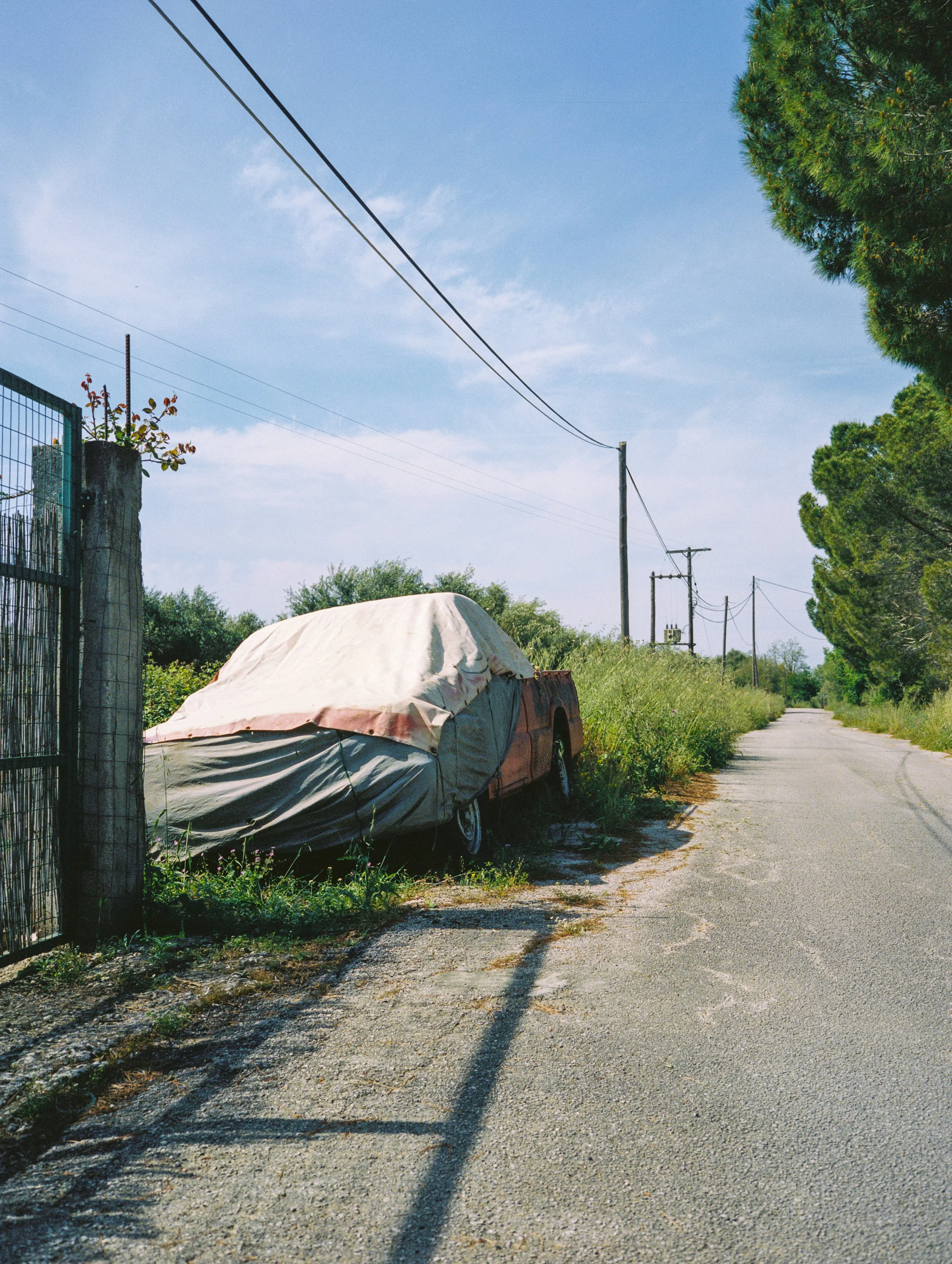 A gravel pathway beside a fence with a tree on the right, a parked car covered with a tarp, and power lines overhead against a clear blue sky.