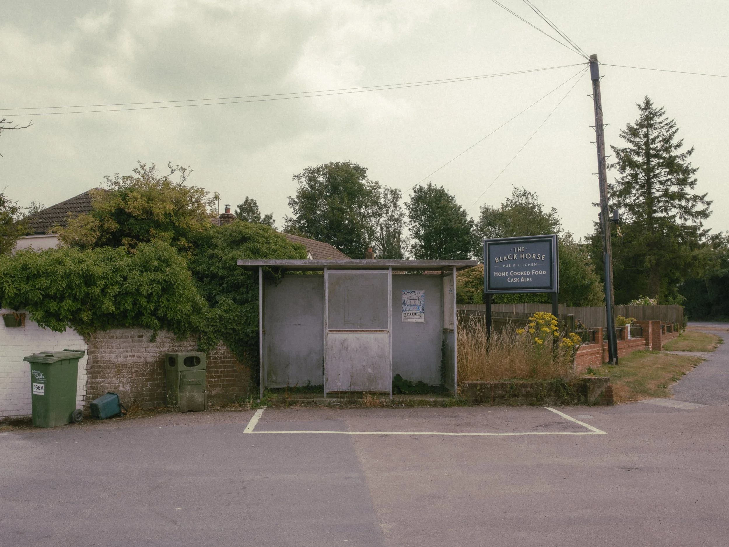 Empty parking spot in front of a bus shelter with a sign for The Black Horse pub and kitchen, surrounded by greenery and trees under an overcast sky.