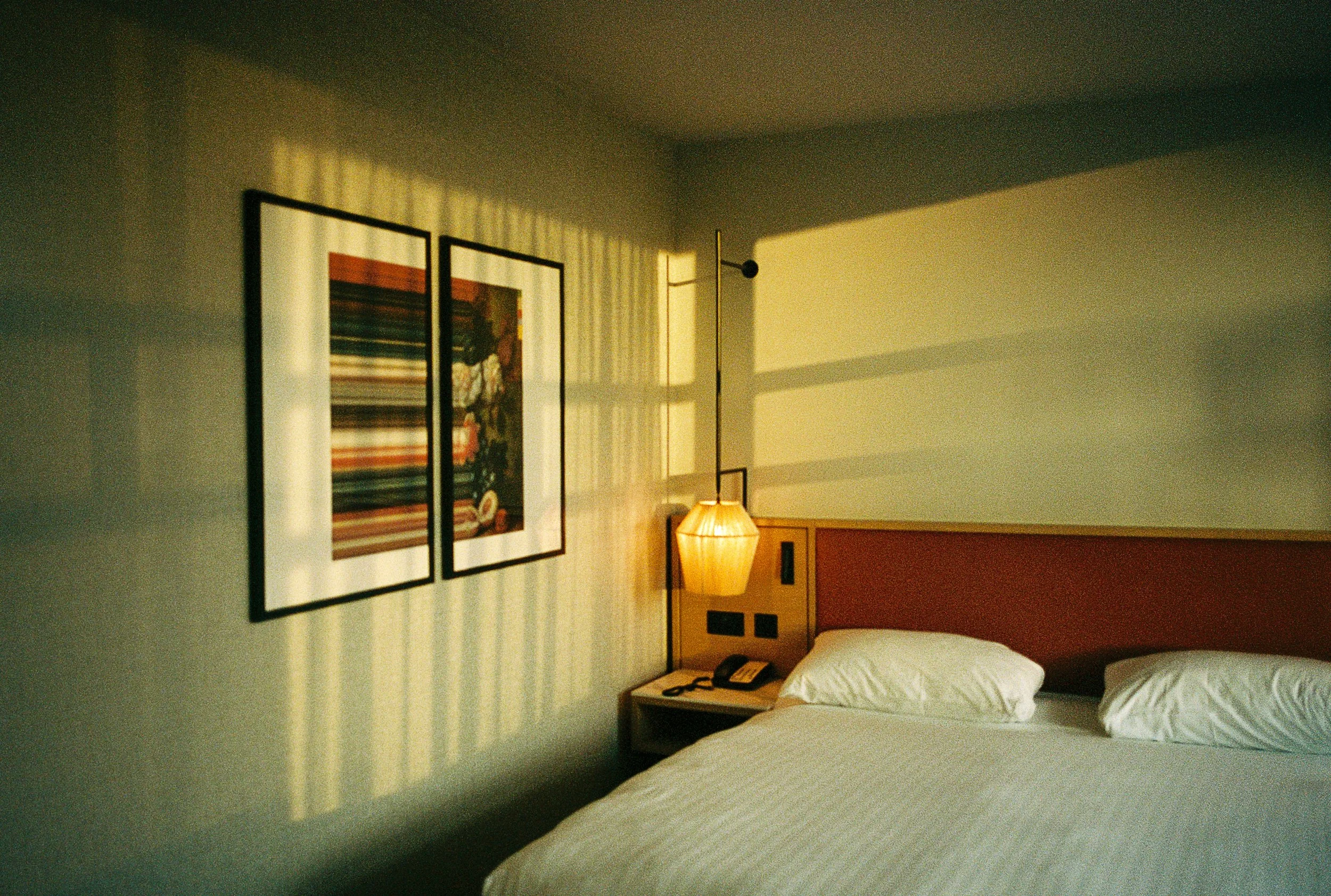A bedroom with two beds, a wooden headboard, a nightstand with a telephone, a table lamp, and hanging wall art featuring abstract patterns, illuminated by sunlight casting shadows on the wall.