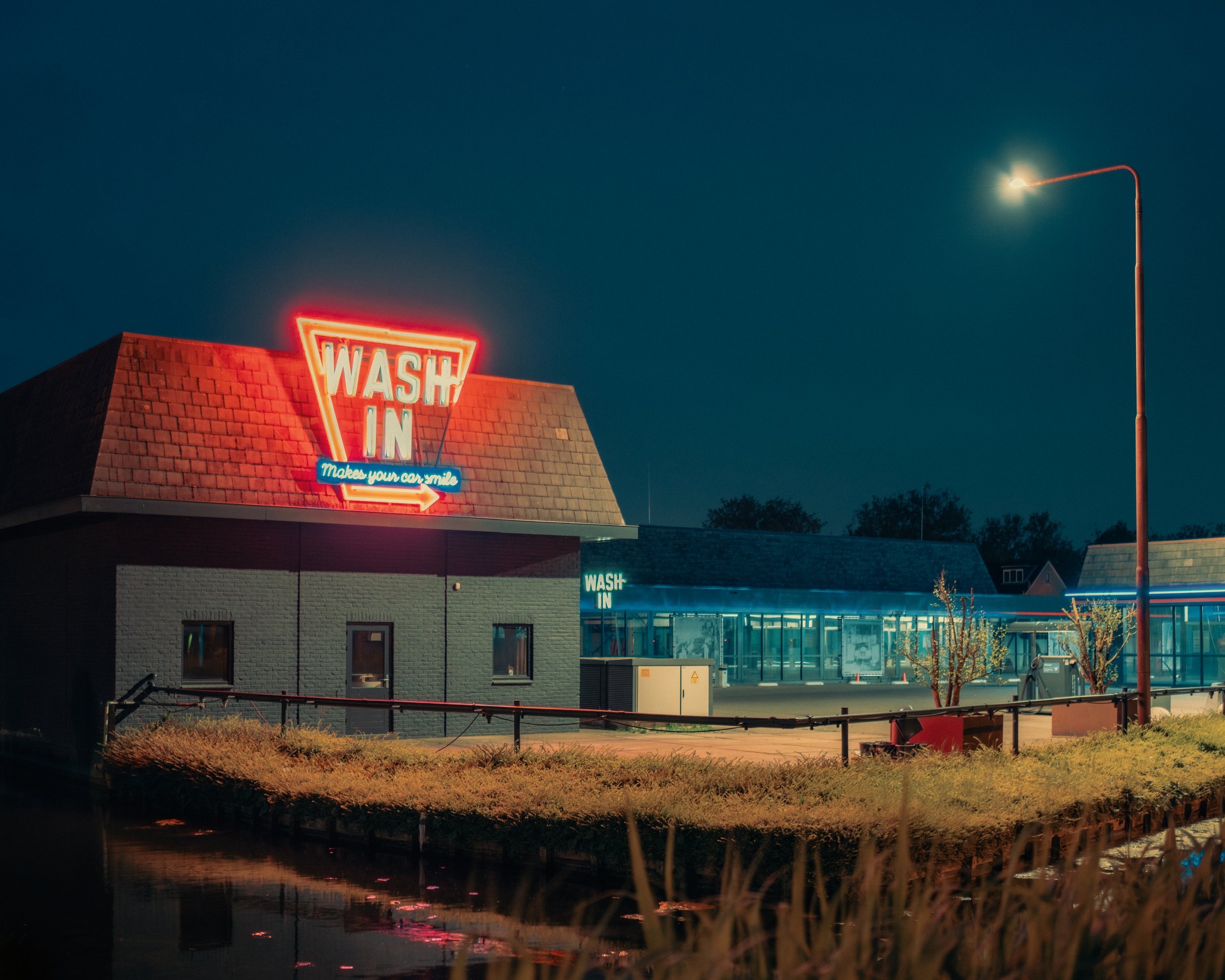 Nighttime scene of a building with a neon sign that reads 'WASH IN' and a small arrow, with city streetlights and a tall streetlamp illuminating the area, and trees and business signs visible in the background.