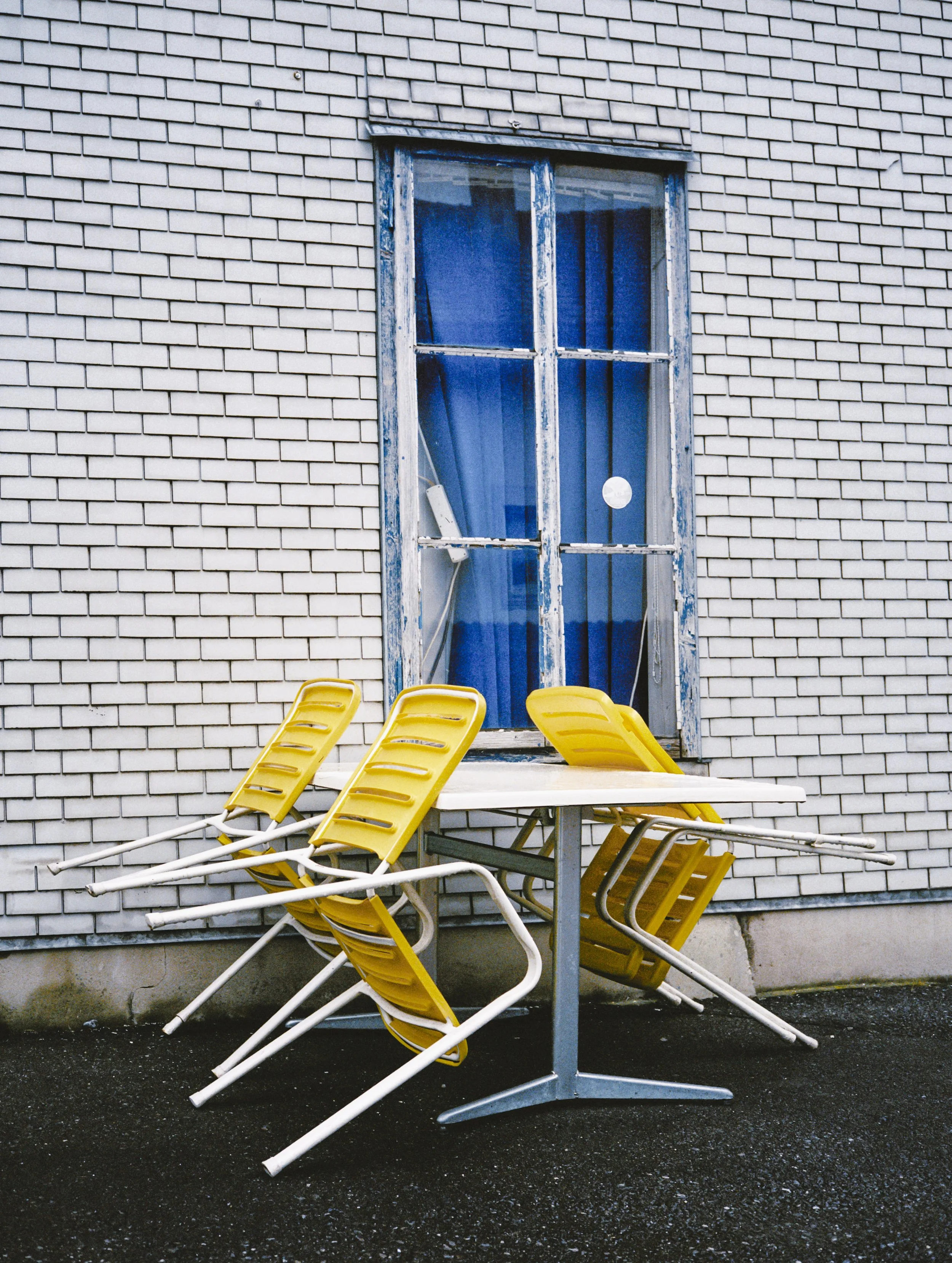 Stack of yellow and white lounge chairs knocked over and leaning against a detached table with a window in a grey brick building in the background.