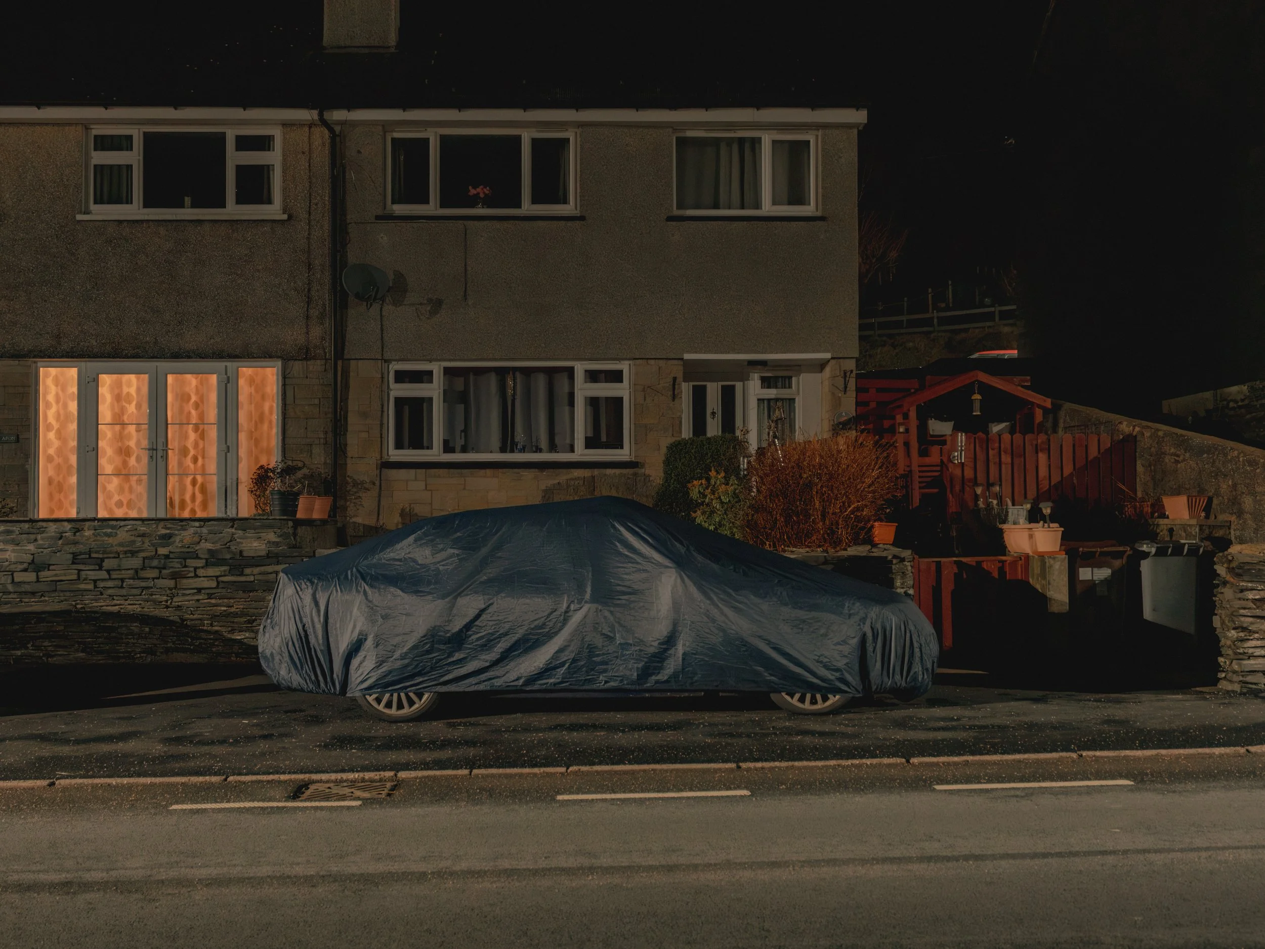 A car parked on the street at night, covered with a gray protective car cover, with a building in the background featuring windows, a satellite dish, a red fence, and outdoor decorations.