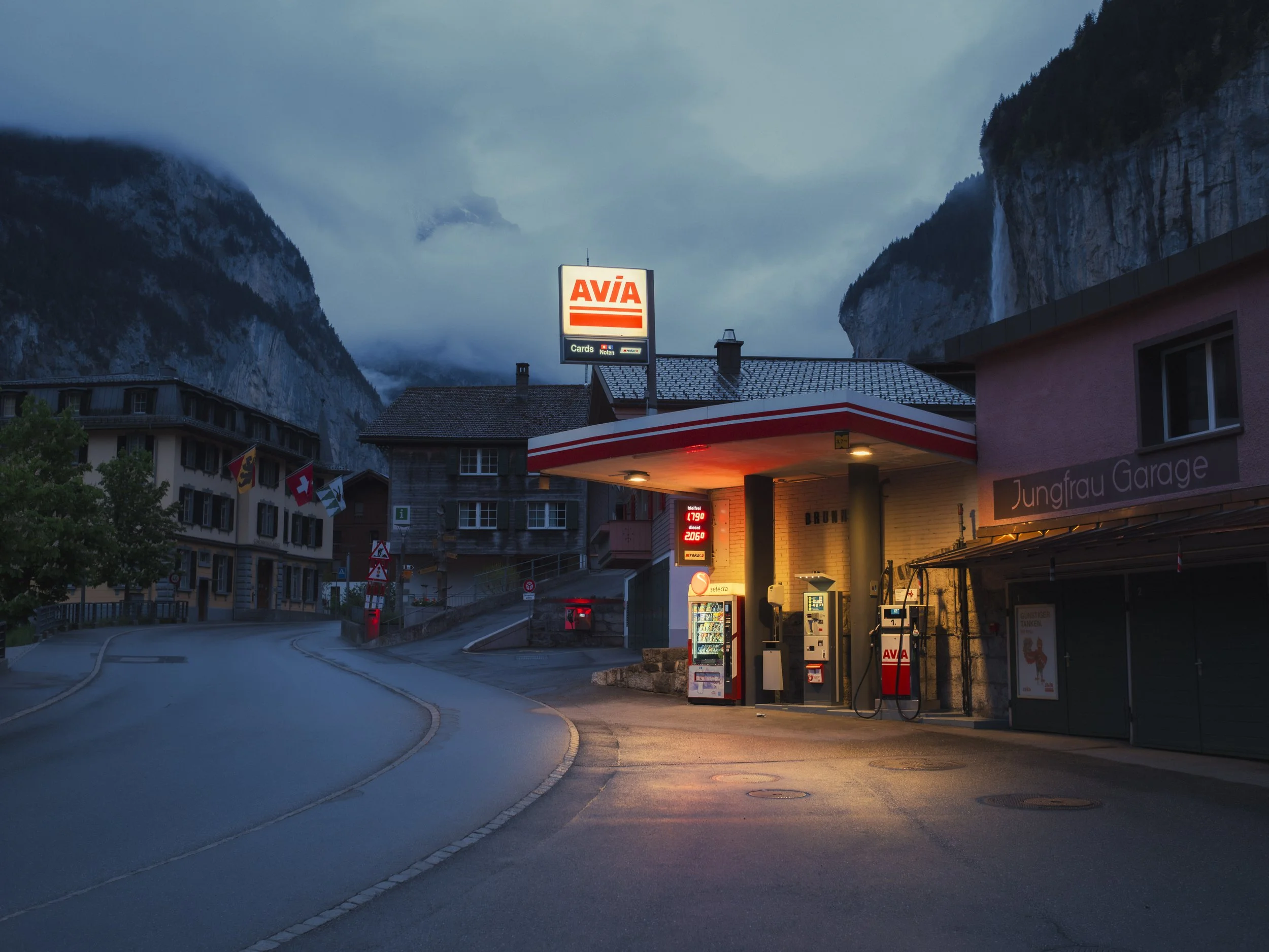 A gas station at dusk with a mountain range and cloudy sky in the background, displaying illuminated signs and fuel pumps under a sheltered canopy.