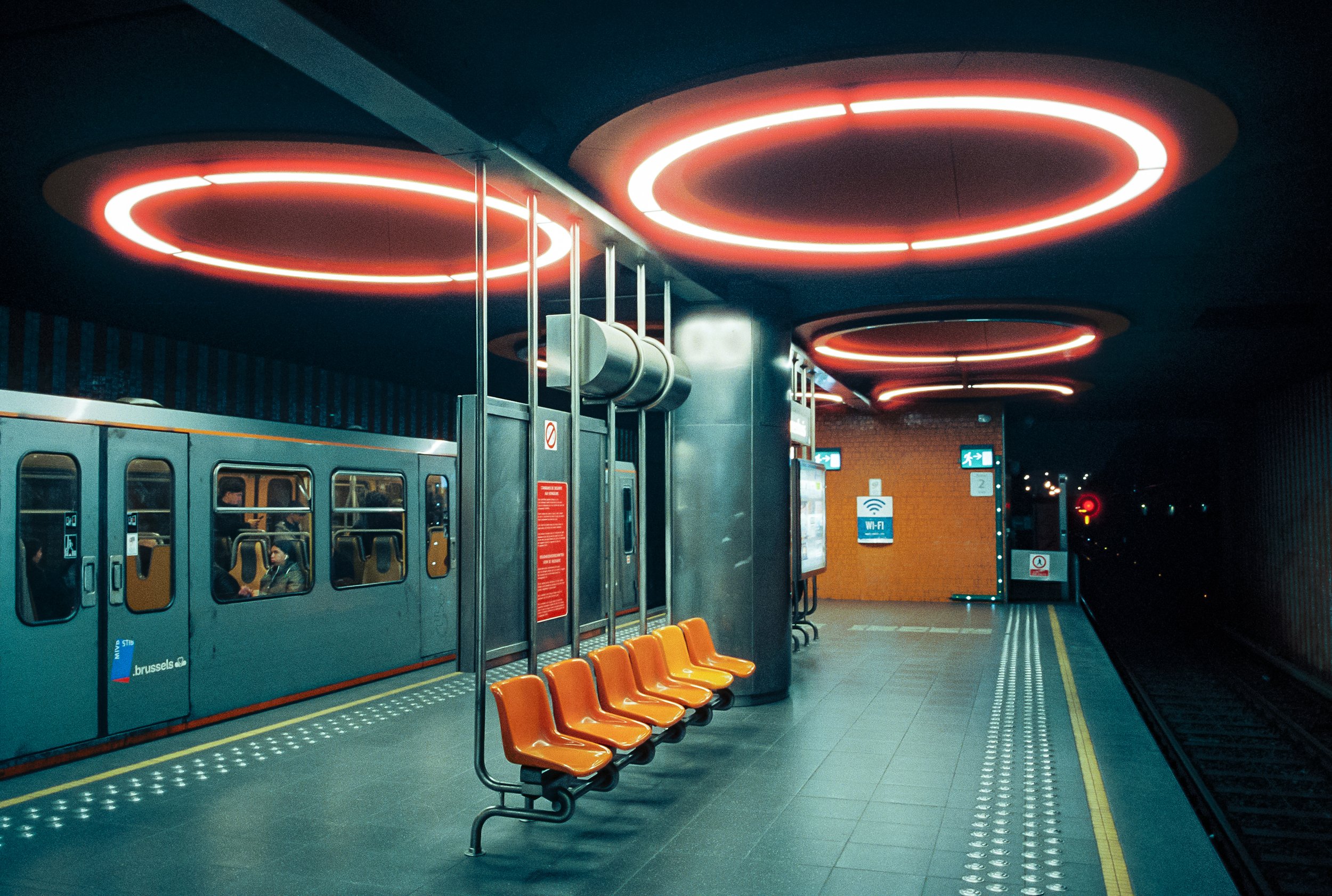 An empty train station platform at night with orange chairs, illuminated red circular ceiling lights, and a train with passengers inside.