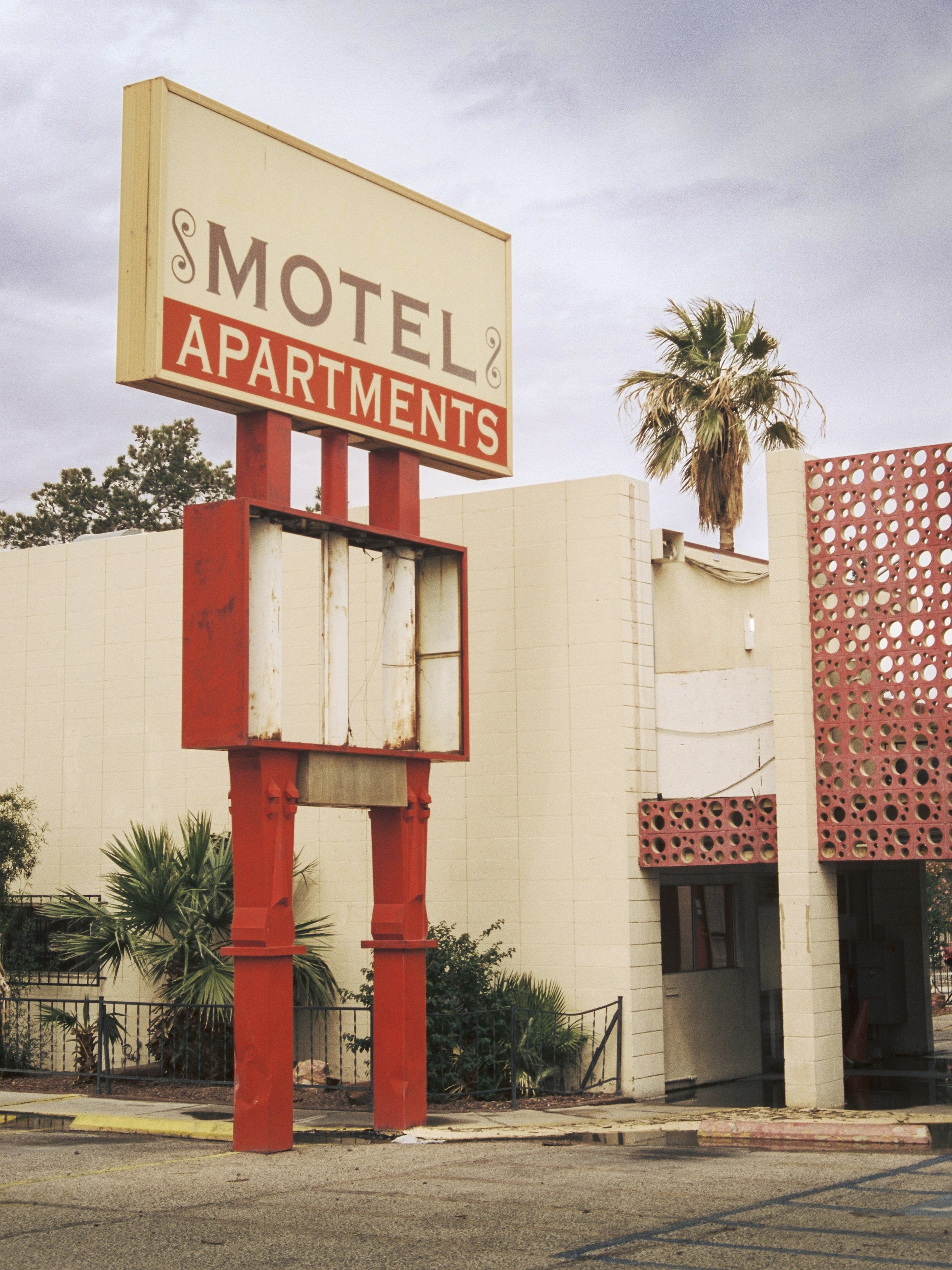An outdoor scene showing an empty sign frame in front of a beige building with a decorative pink lattice panel, a tall palm tree, and some bushes, under a cloudy sky.