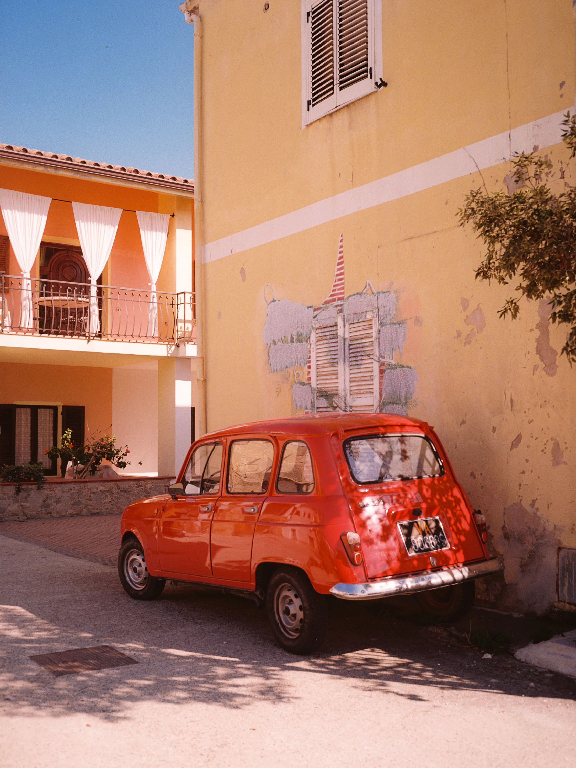 A small orange vintage car parked on a street near a beige building with a mural of a fish and a window with shutters.