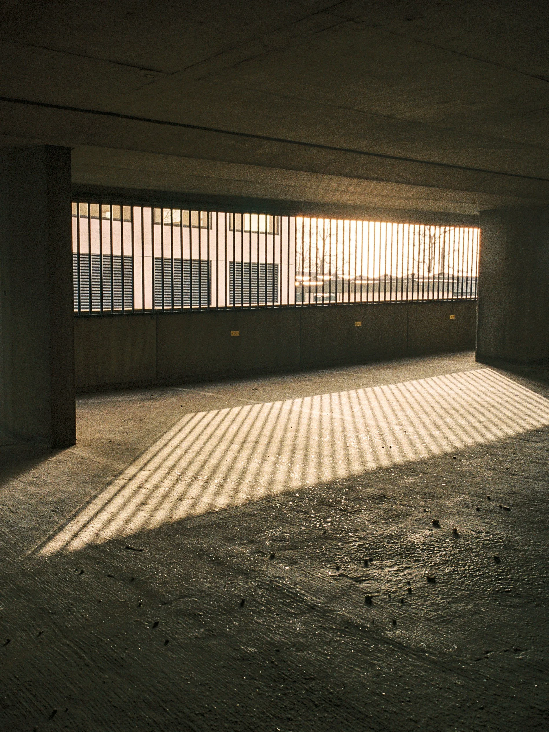 The photo shows the shadow of metal bars cast on the ground, with a building with window shutters in the background, and sunlight coming through the bars.