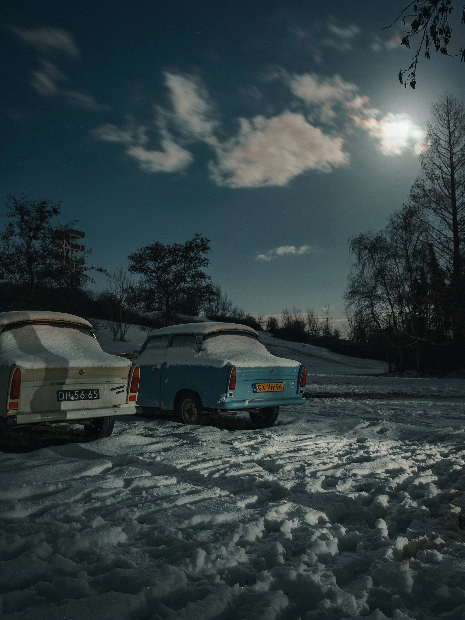 Snow-covered vintage cars parked in a snowy lot at night with a bright moon in the sky, bare trees, and a rural landscape.