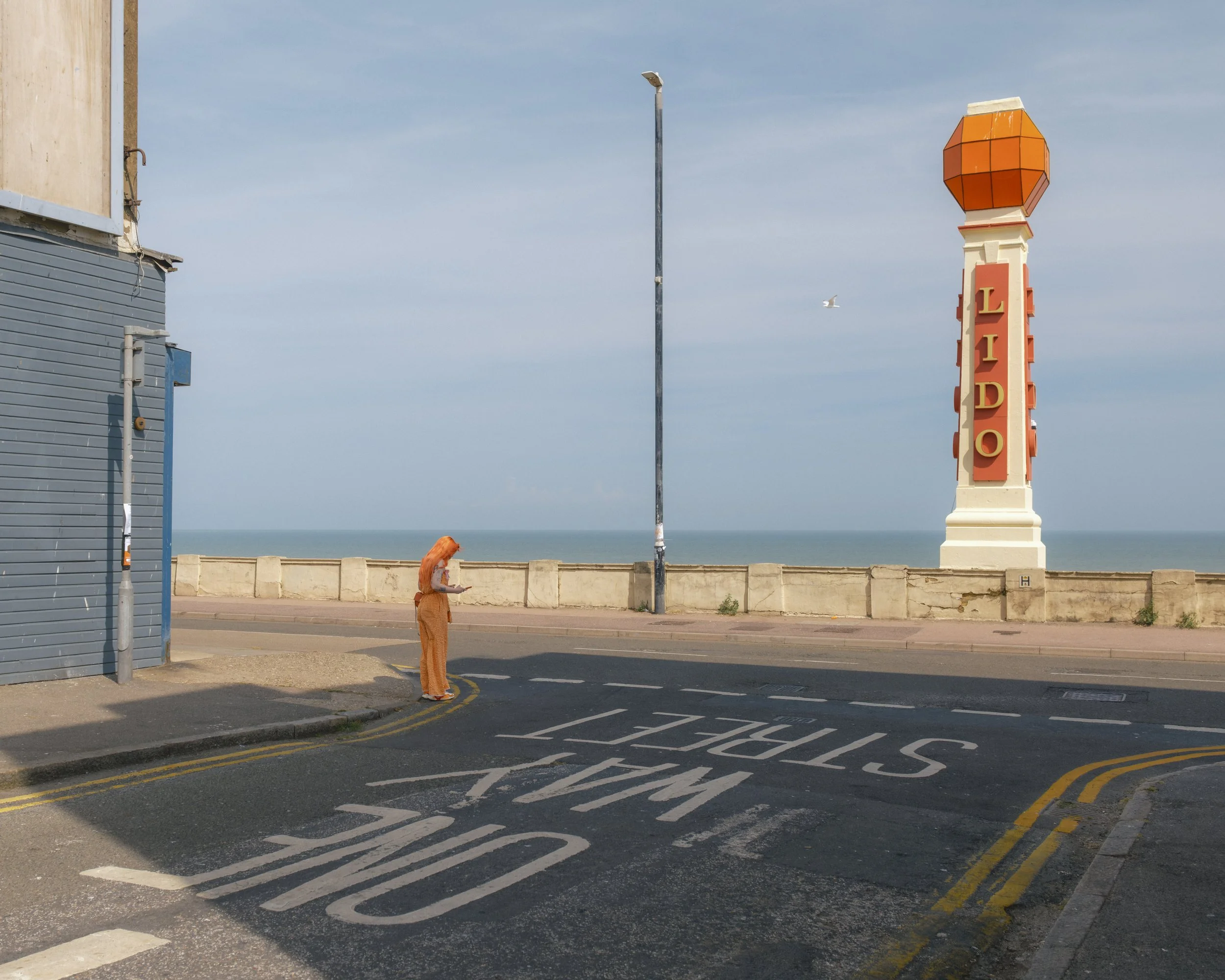 A woman with orange hair and orange pants standing on a sidewalk looking at her phone, near a street with white and yellow road markings, next to a blue building and a tall orange and white sign with the word 'LIDO' against a blue sky with few clouds