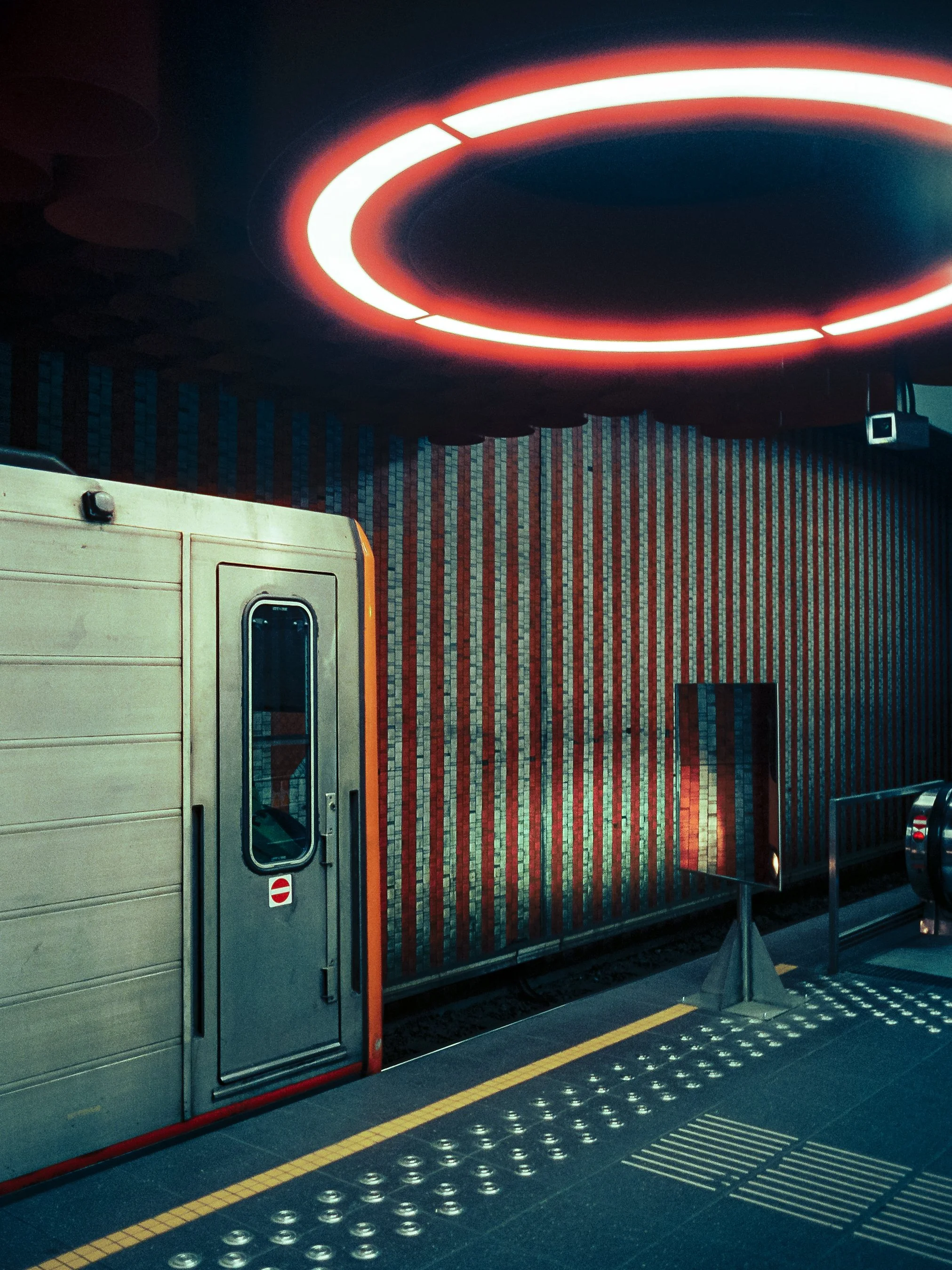 Inside a bus or train station, there is a platform with a yellow tactile strip and silver circular sensors for visually impaired individuals. A section of the station's wall, which has a red, white, and blue mosaic design, is visible along with a par