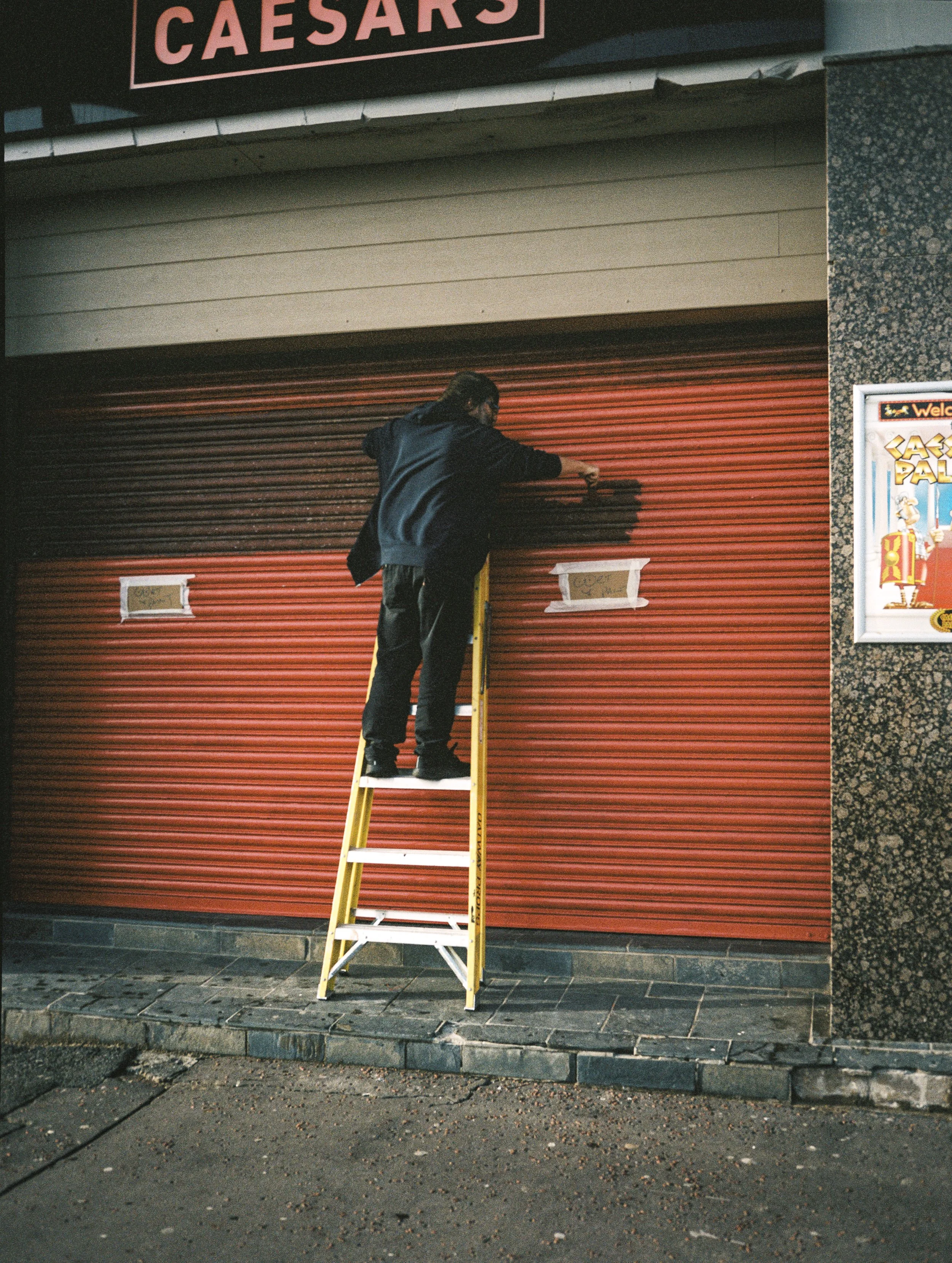 A person is standing on a yellow stepladder in front of a closed red metal shutter. The person appears to be painting or working on the shutter. There is a sign above with the word 'CAESARS' and a poster on the wall to the right. The scene is outdoor