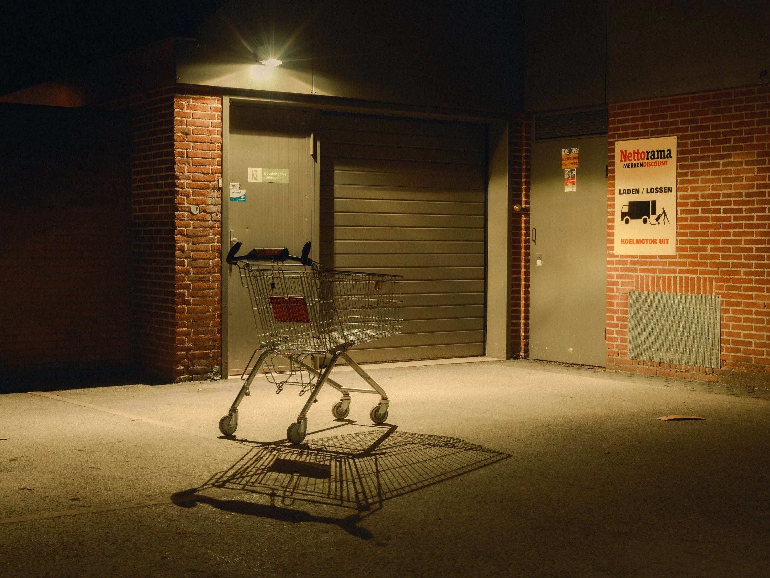 Empty shopping cart casting shadow on pavement outside a brick building at night, illuminated by exterior light.