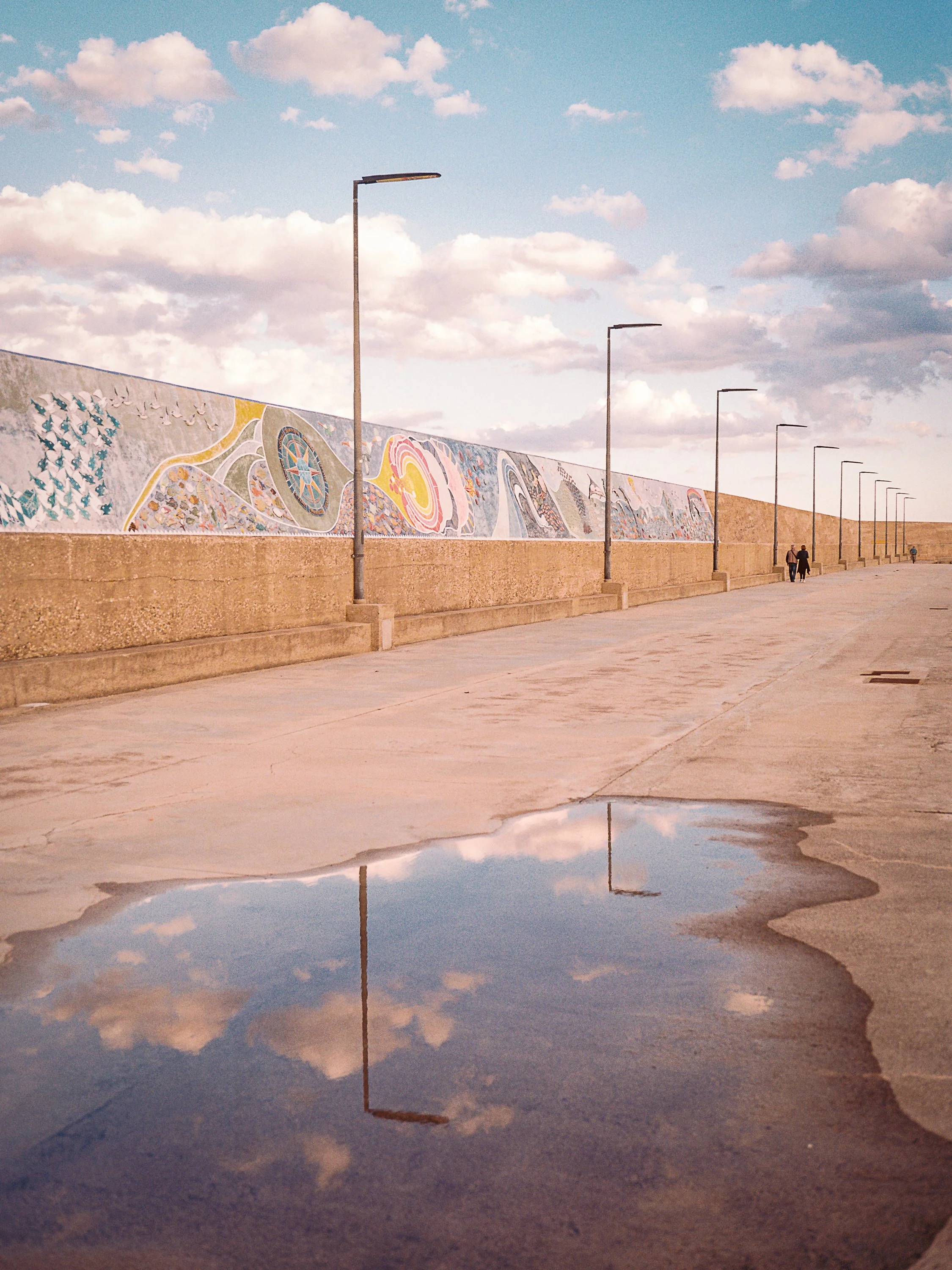 Street with sidewalk, decorative wall with mural art, streetlights, cloudy sky, and reflections of clouds in a puddle on the ground.