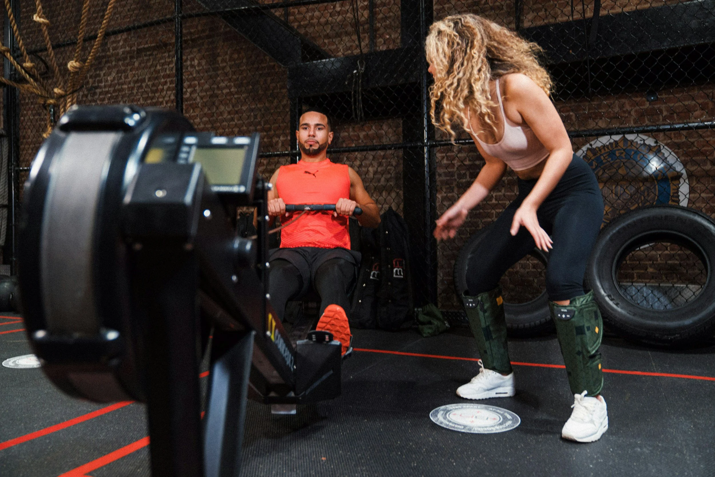 A woman in workout attire adjusting her socks while a man in a fitness gym sits on a bench holding a resistance band, with gym equipment and a brick wall in the background.