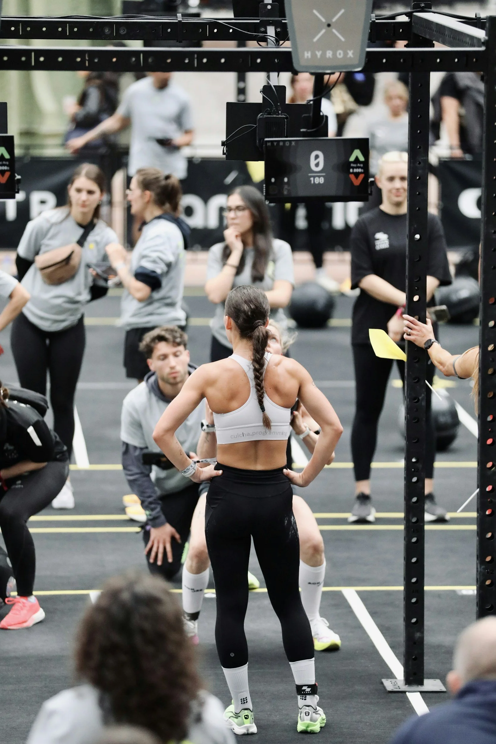A woman with a tattoo on her back, wearing a white sports bra and black leggings, standing in front of a group of people in an indoor gym or fitness space. Several people are seated or standing in the background, some talking and others looking at the woman.