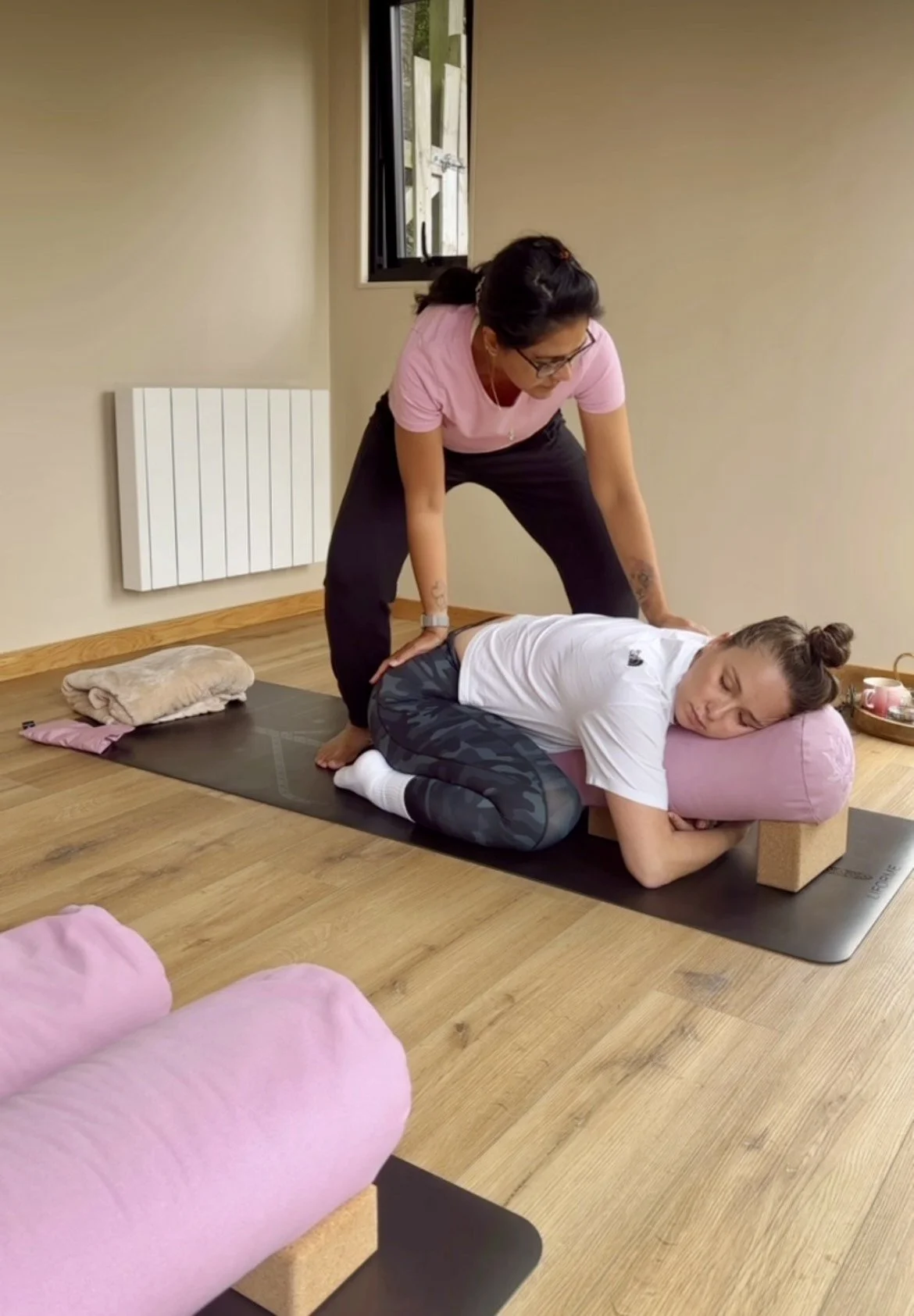 A woman practicing yoga on a black mat, resting her head on a pink pillow, with an instructor assisting her in a kneeling position in a room with wooden floors, beige walls, and a window.