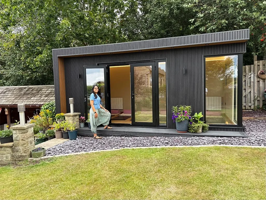 A woman standing on a small deck in front of a modern black tiny house with large glass doors and windows, surrounded by potted plants and a well-maintained lawn.