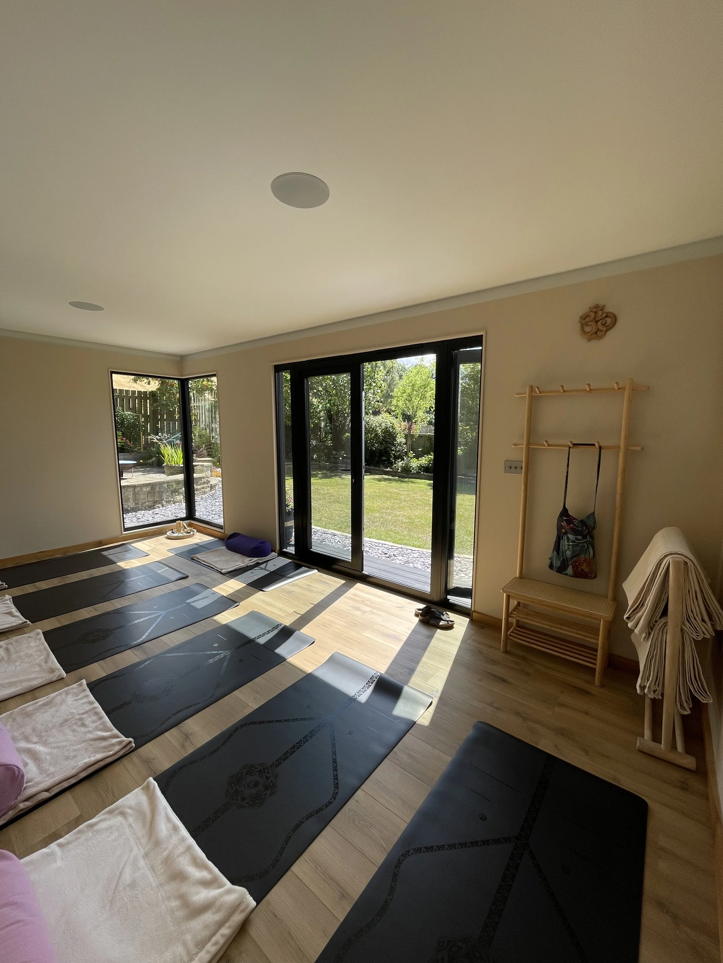 A sunlit yoga or meditation room with yoga mats laid out on a wooden floor, large glass sliding doors opening to a lush garden, and a wooden coat rack with a colorful bag hanging on it.