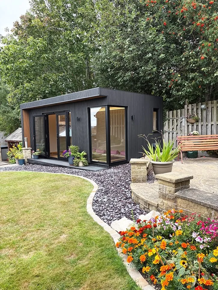 Black modern tiny house with large glass sliding doors in a garden, surrounded by trees, flowers, and a stone pathway.