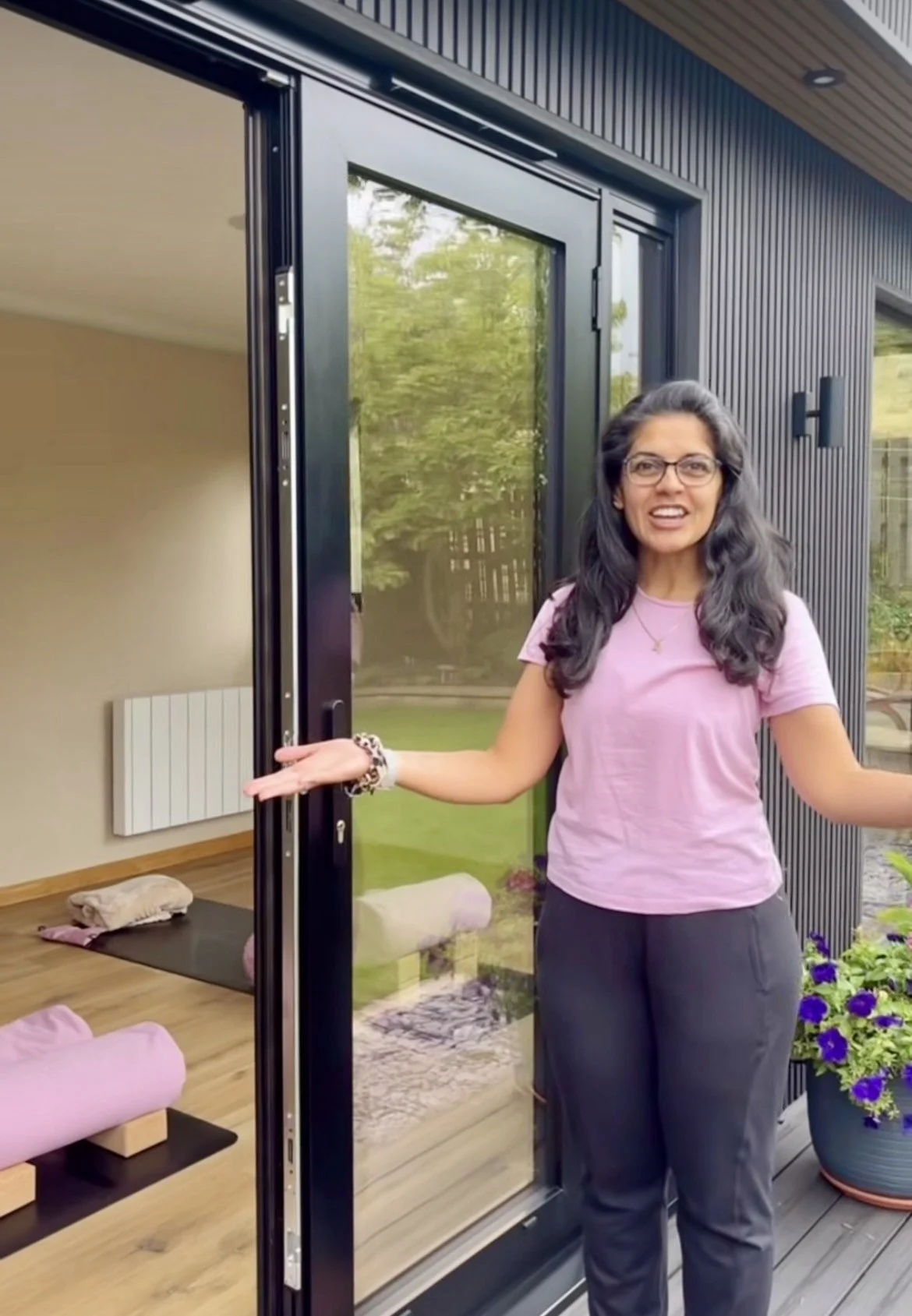 A woman standing outside a house opening a sliding glass door, indoor room visible with a yoga mat, pillows, and a blanket, and an outdoor garden with green grass and plants.