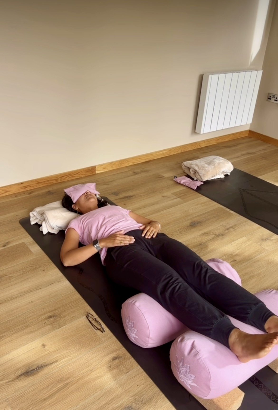 A woman is lying on a yoga mat with a small pillow under her head, appearing to be asleep or resting. She is dressed in a pink shirt and black pants, with a cloth on her forehead and a watch on her left wrist. There is a pillow and a blanket beside her, and another yoga mat and pillow are visible nearby in a room with wooden flooring and a radiator.